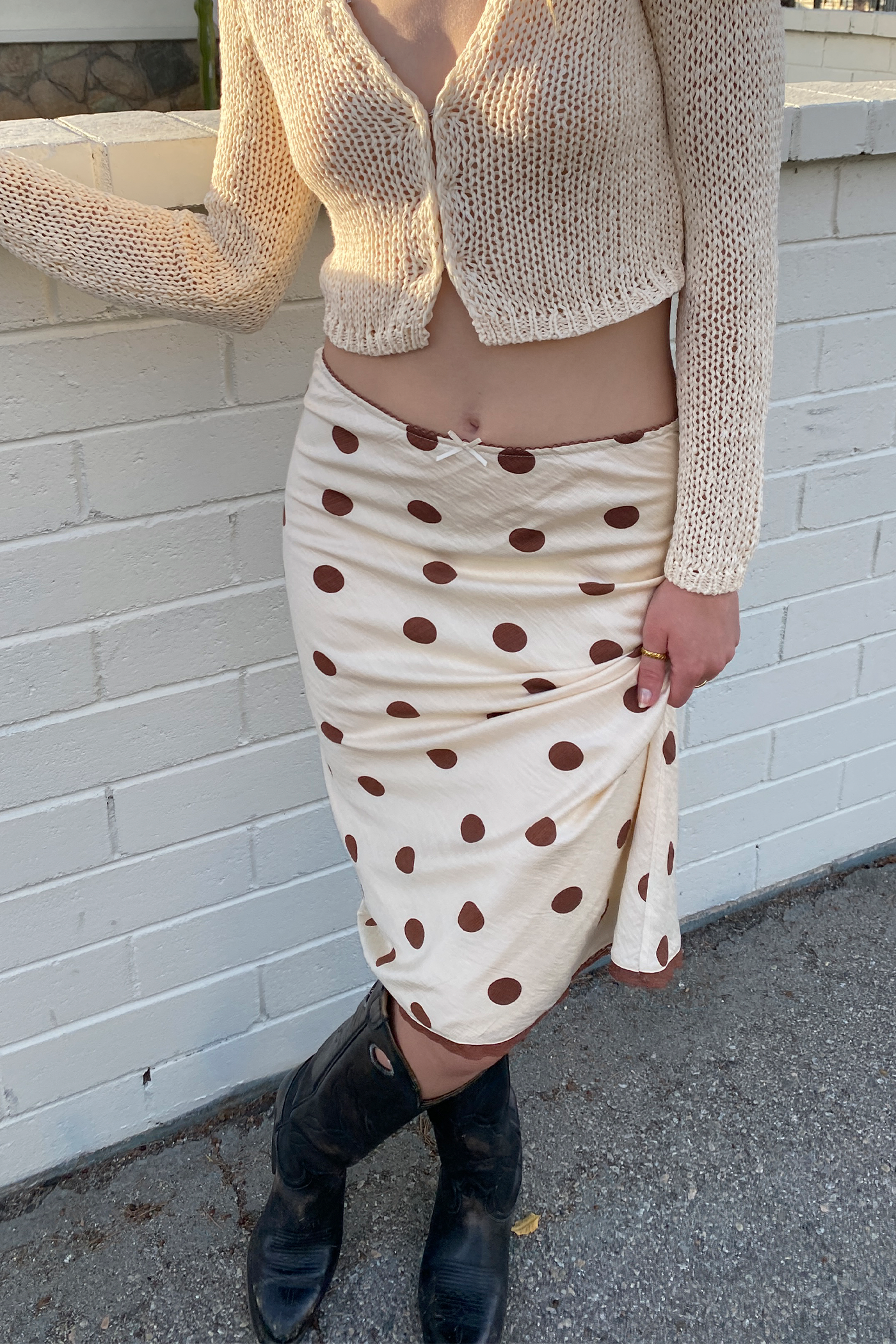 Person wearing a beige knit cardigan and polka dot skirt against a white brick wall.