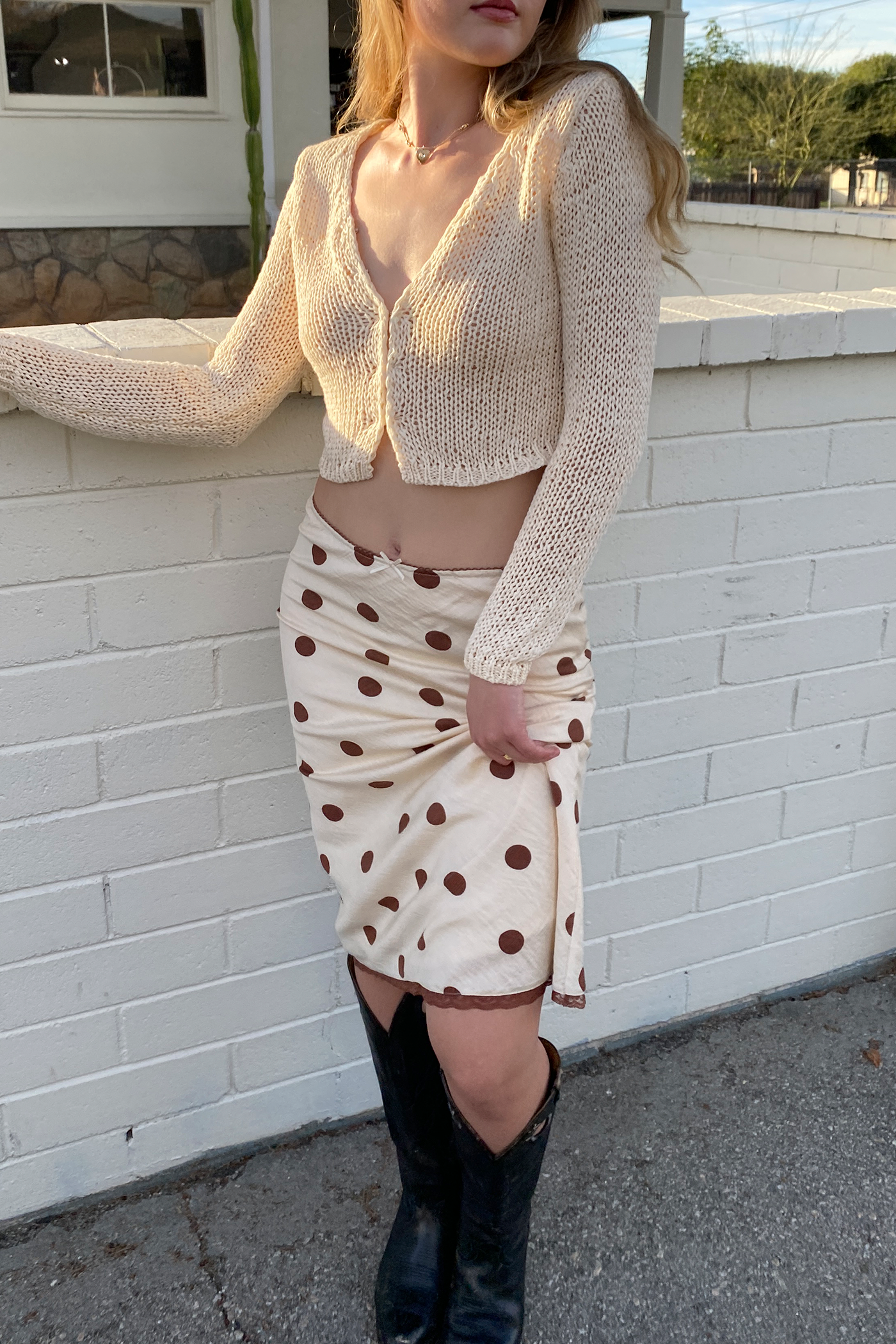 Woman wearing a beige cardigan, polka dot skirt, and black boots against a white brick wall.