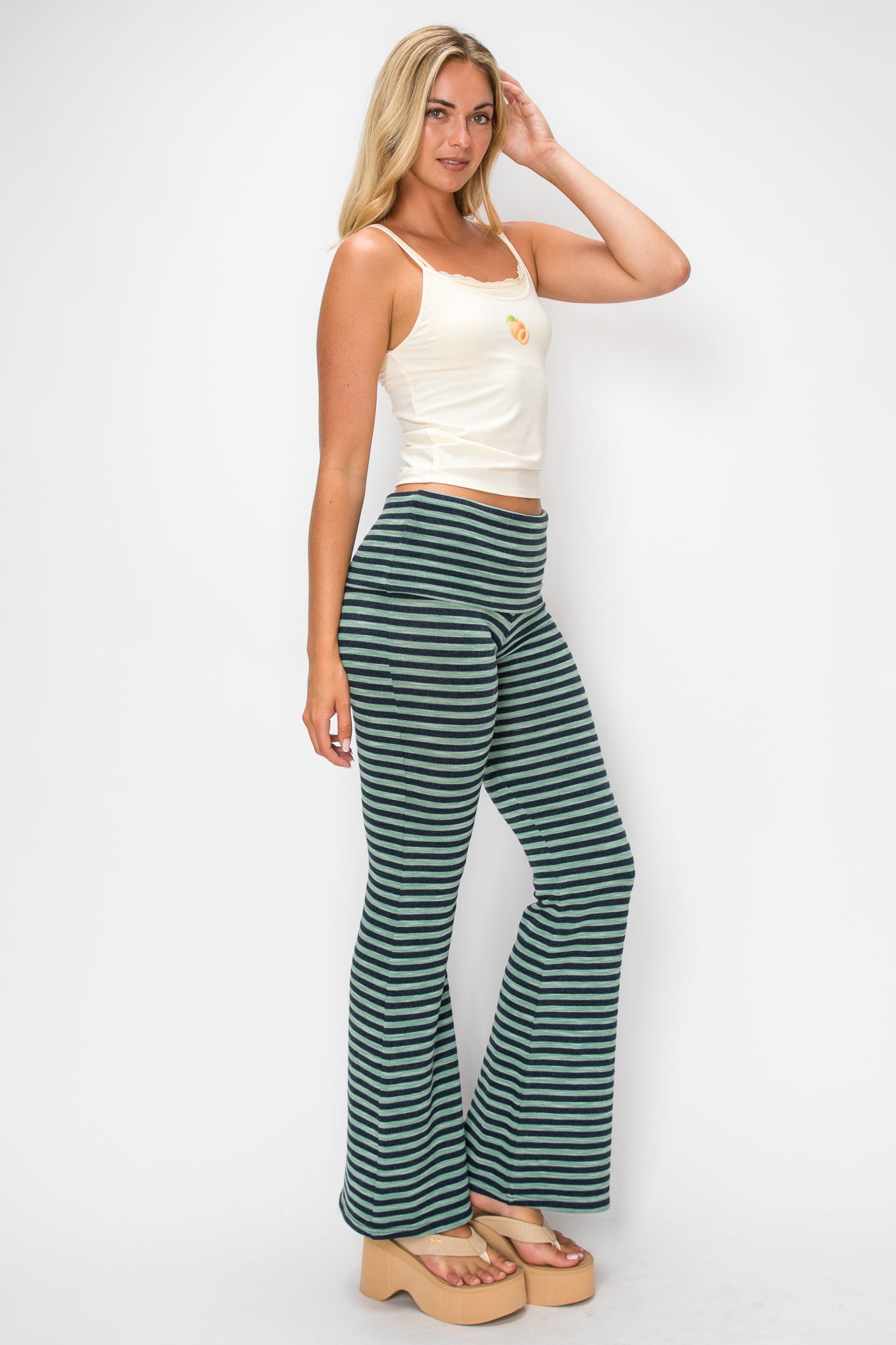 Woman wearing a white tank top with a fruit design and green pants against a plain background