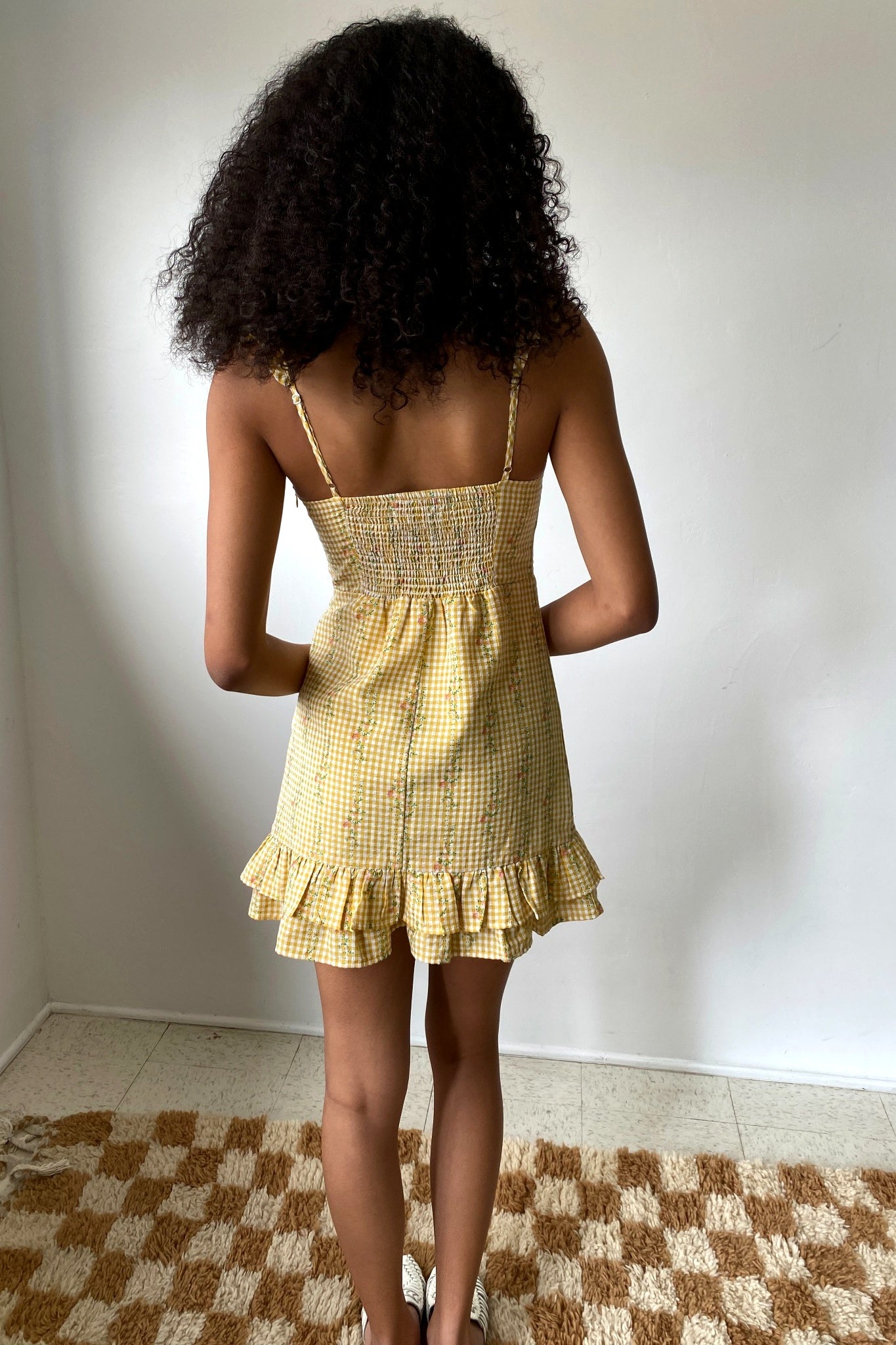 Person wearing a yellow checkered dress standing against a white wall.