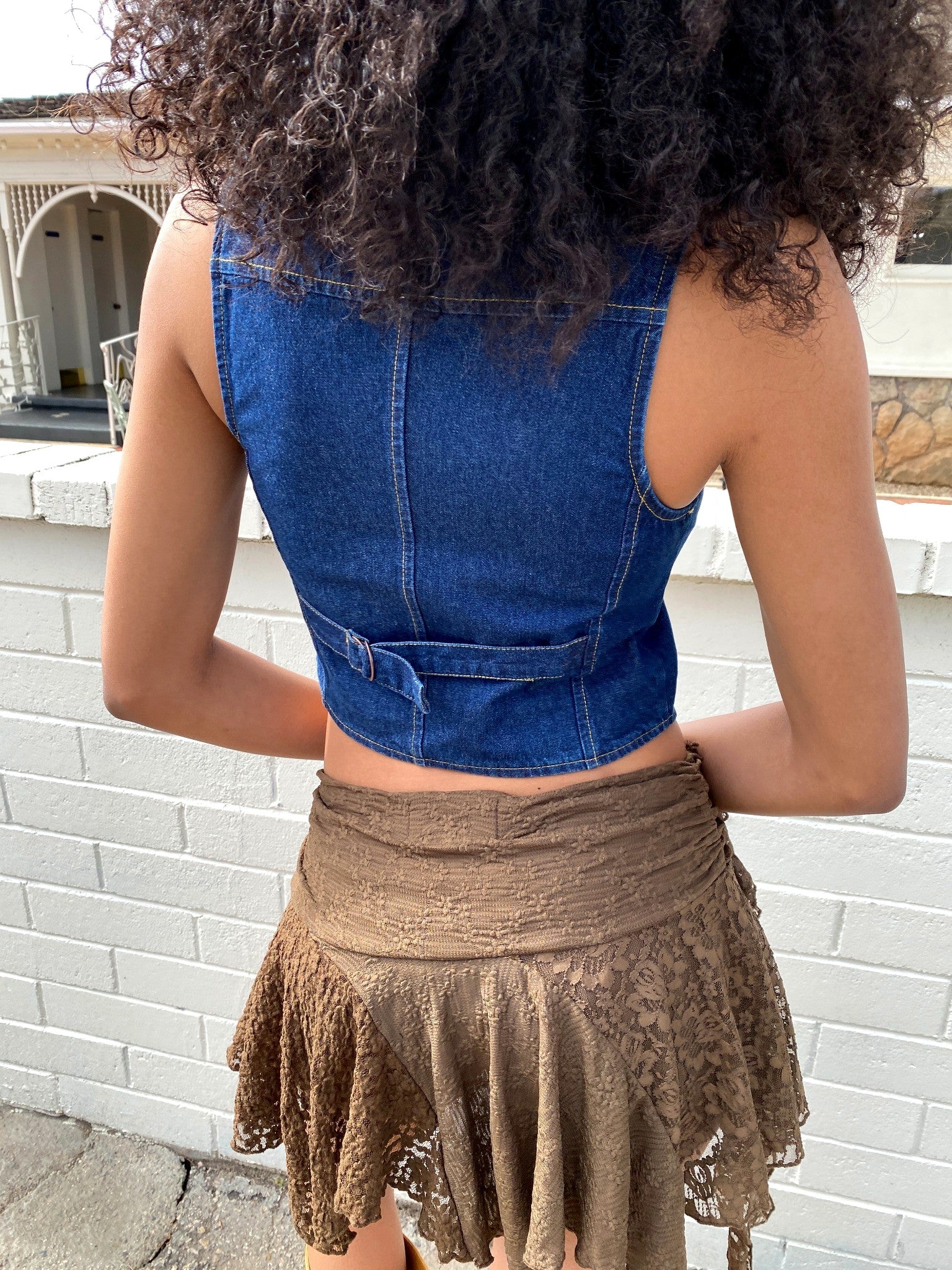 Person wearing a blue denim top and brown lace skirt against a white brick wall.
