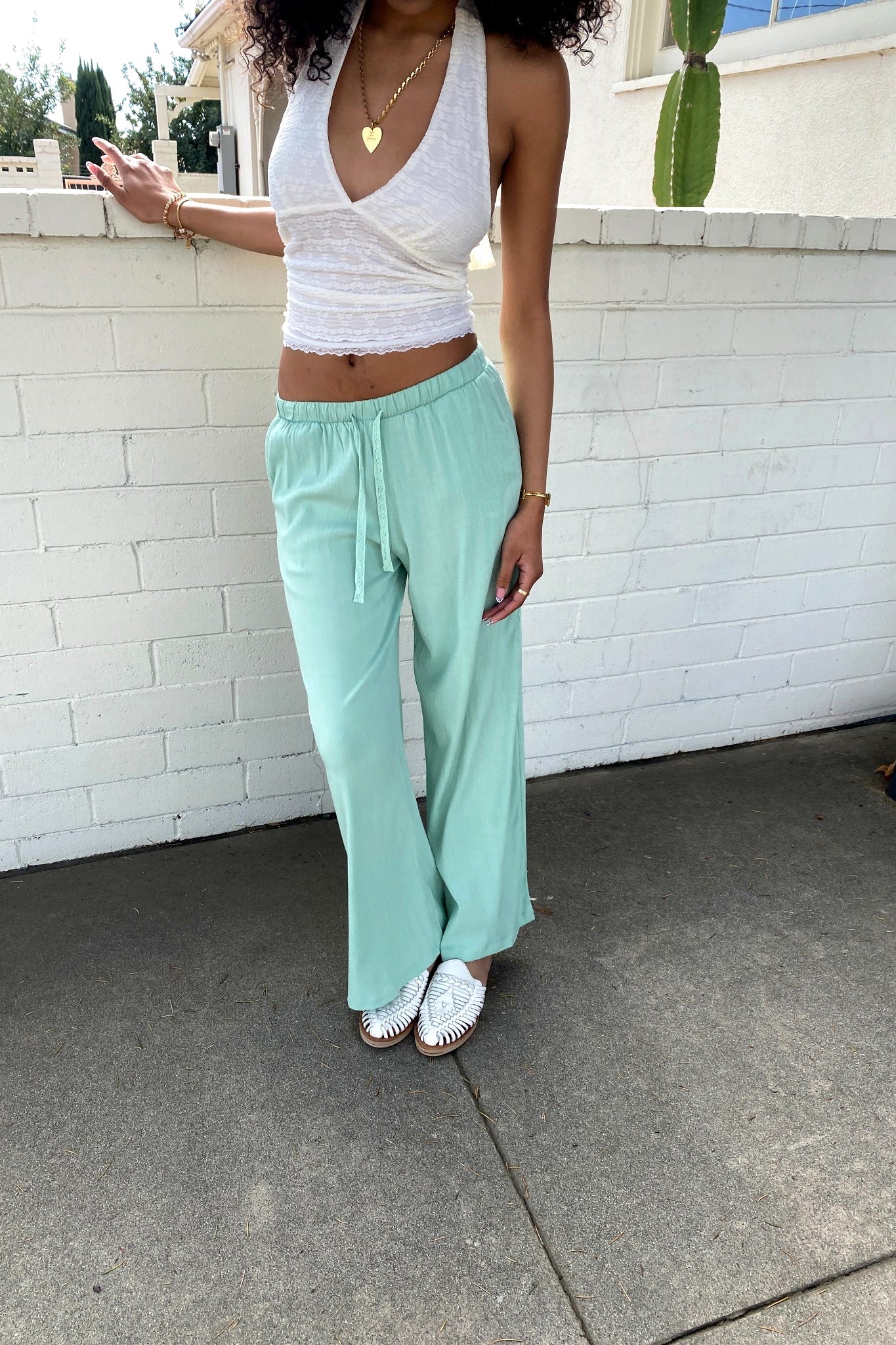 Woman wearing a white top and light green pants standing against a white brick wall.