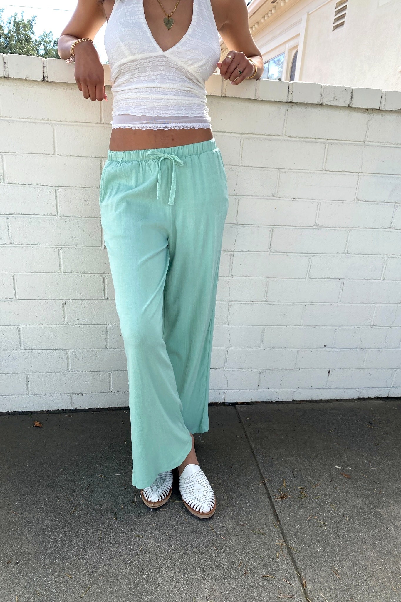 Person wearing a white top and light green pants standing against a white brick wall.