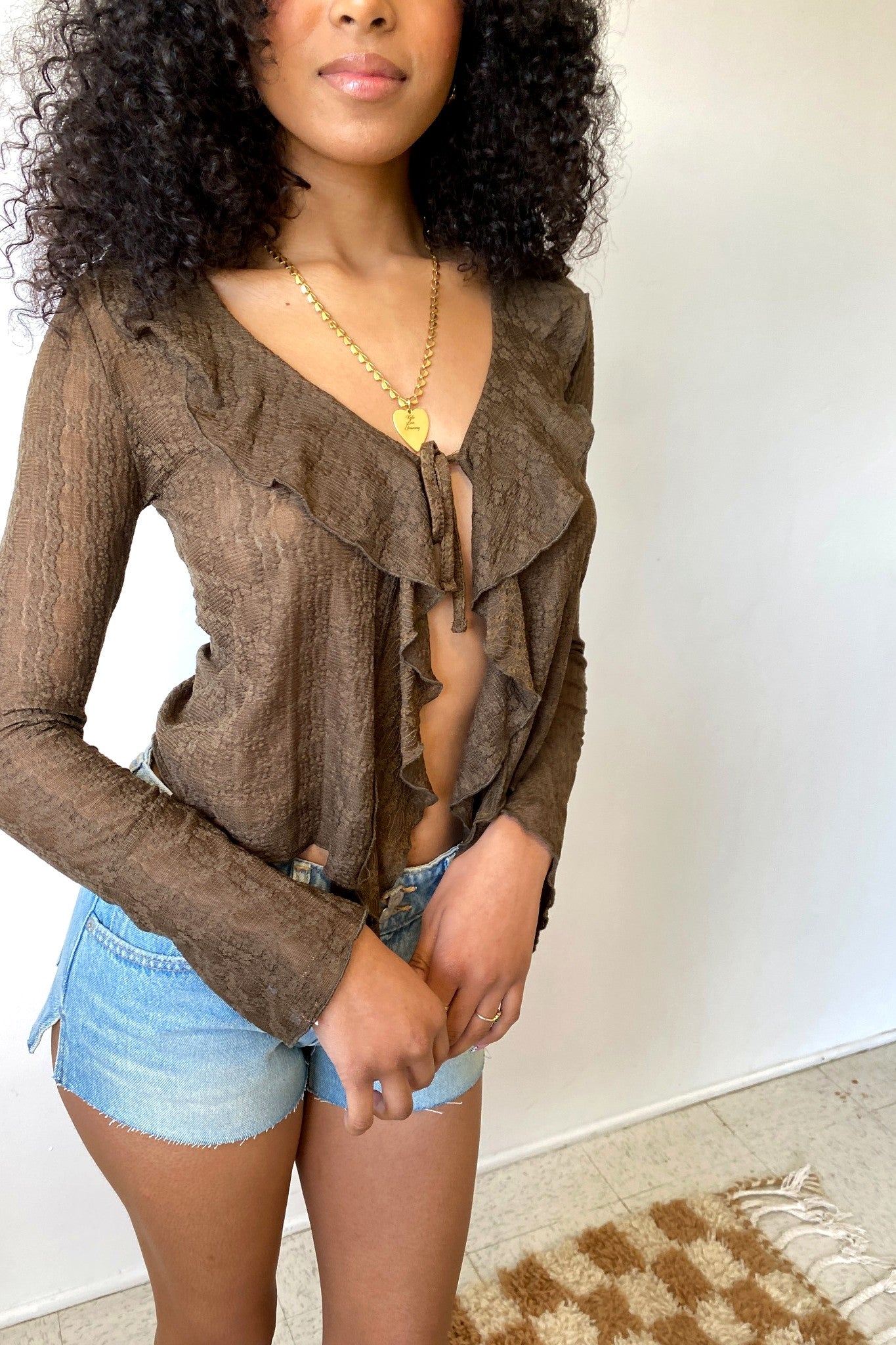 Woman wearing a brown sheer top and denim shorts against a white wall.