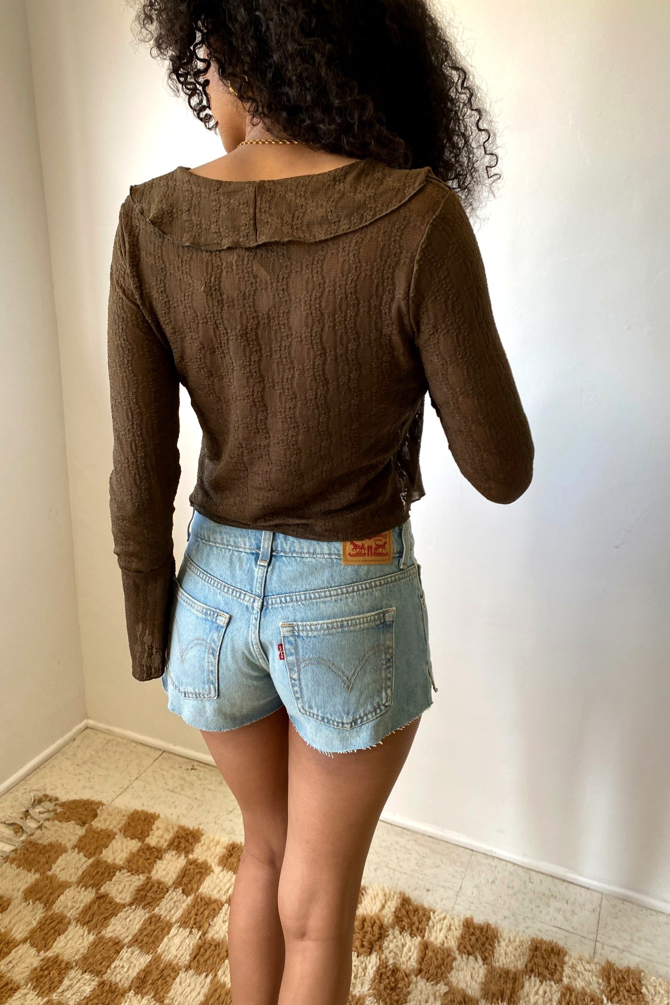 Person wearing a brown long-sleeve top and light blue denim shorts in a room with a checkered floor.