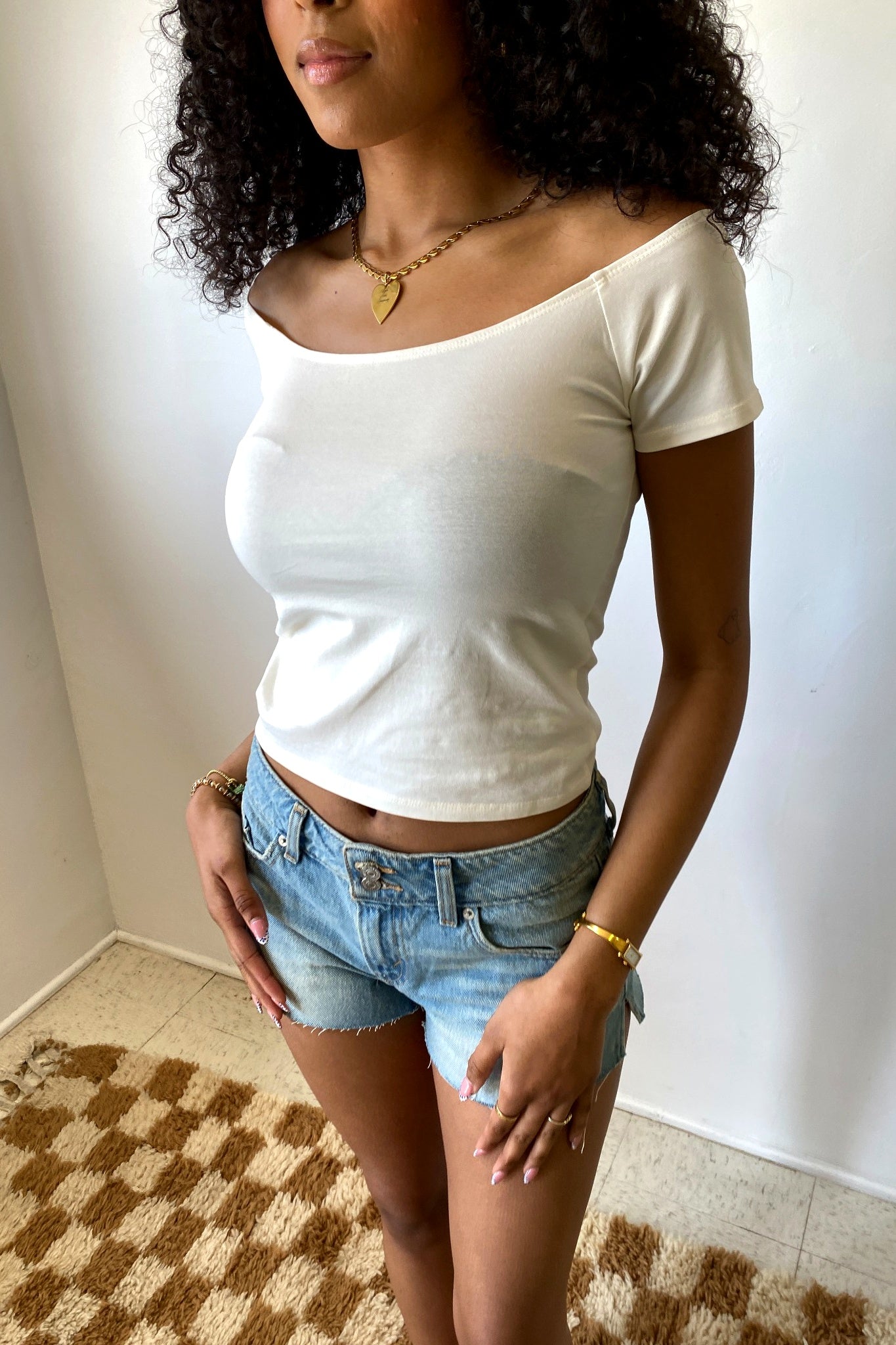 Woman wearing a white top and denim shorts standing against a plain wall.