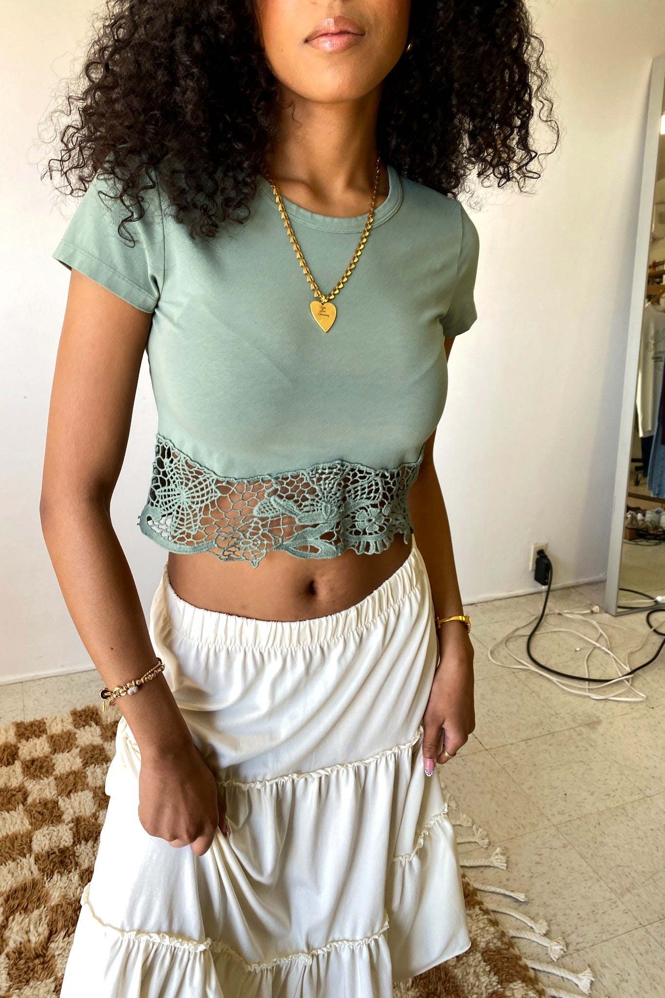Woman wearing a light green top with lace detail and a white skirt indoors.