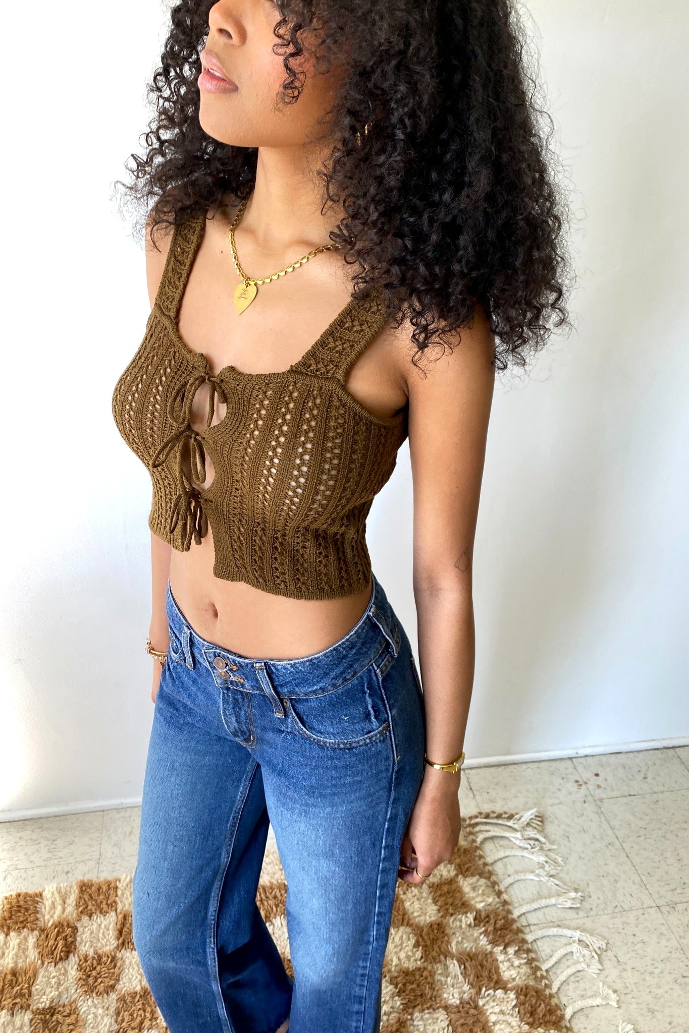 Woman wearing a brown crochet top and blue jeans against a white wall.