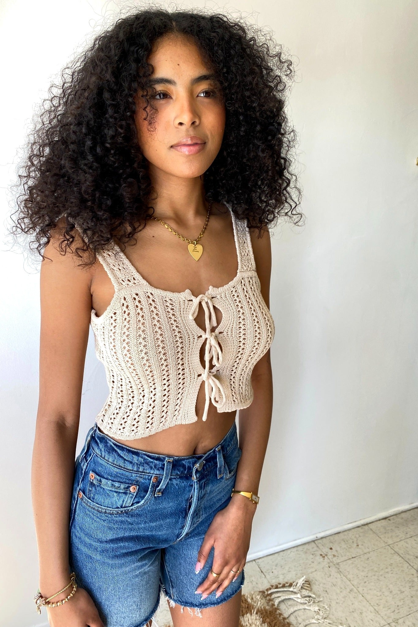 Woman wearing a beige crochet top and blue shorts against a white wall.