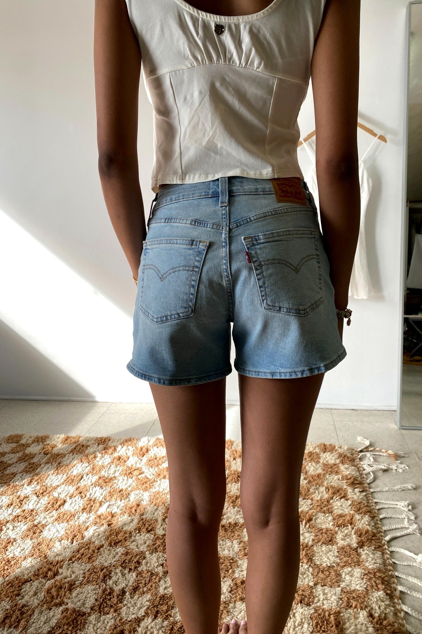 Person wearing a white sleeveless top and blue denim shorts standing on a patterned rug.