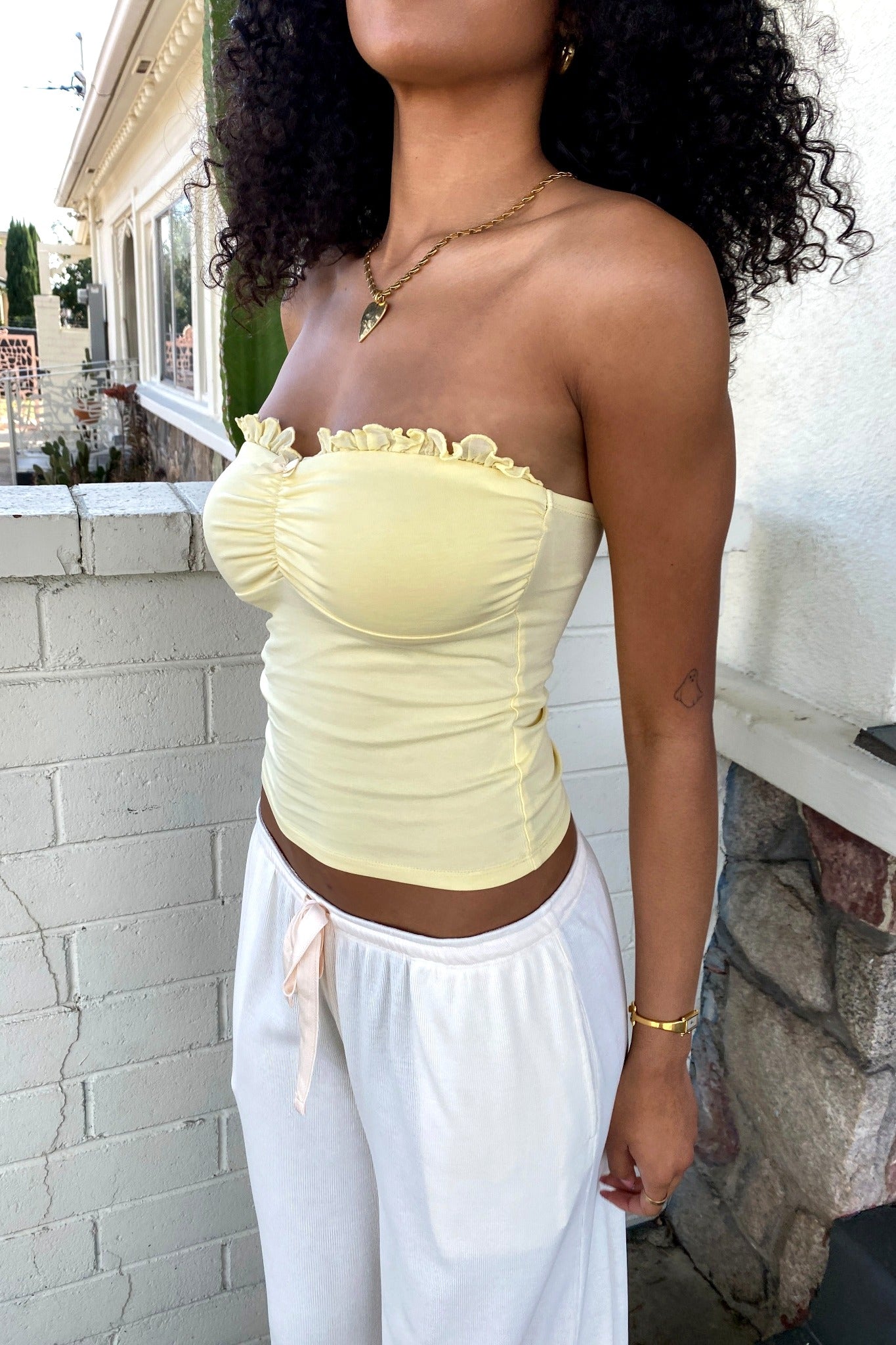 Woman wearing a yellow strapless top and white pants standing against a light-colored wall.