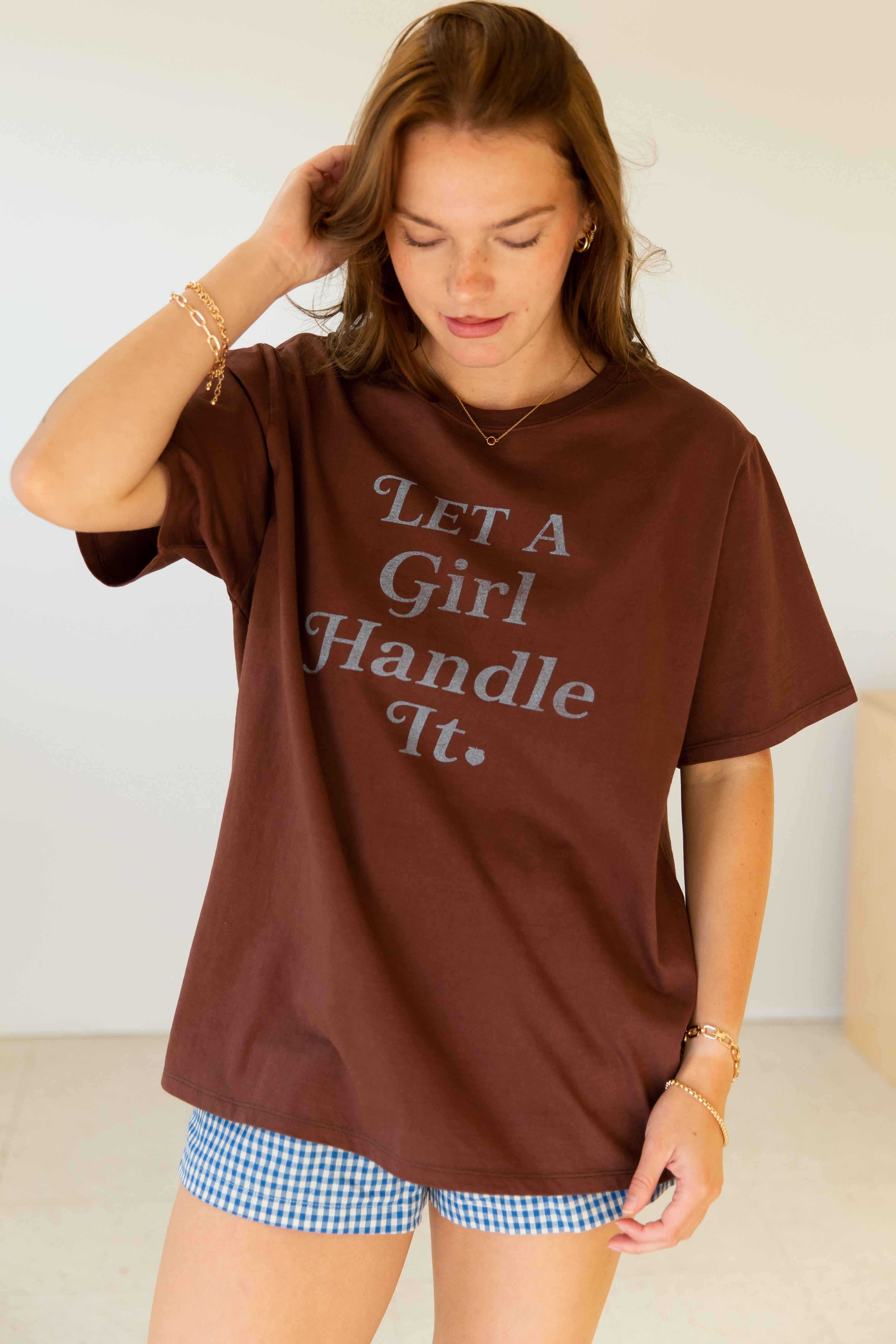 Woman wearing a brown t-shirt with 'Let a Girl Handle It.' text, standing against a plain background.