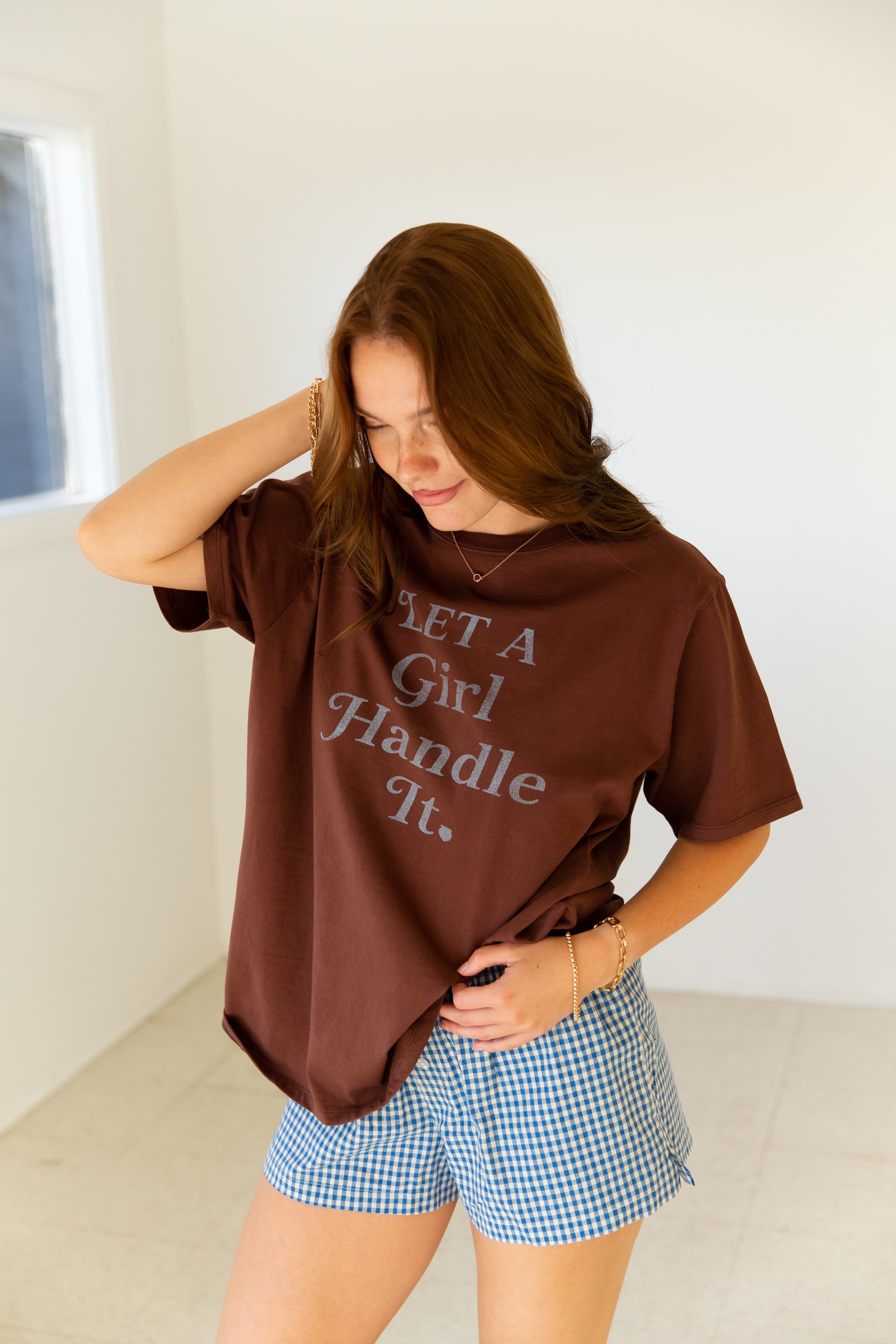 Woman wearing a brown oversized t-shirt with text and blue checkered shorts indoors.