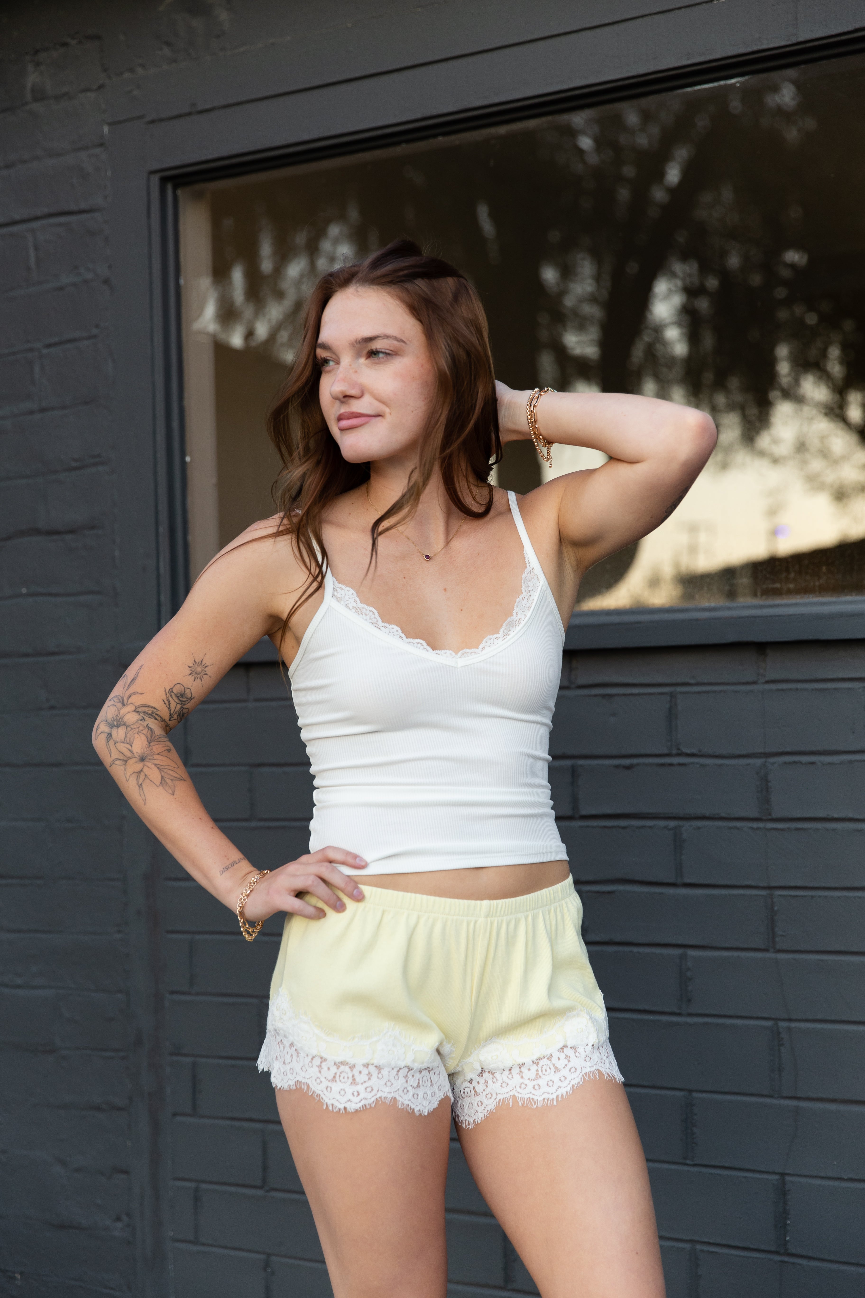 Woman wearing a white tank top and light yellow shorts with lace trim against a dark brick wall.
