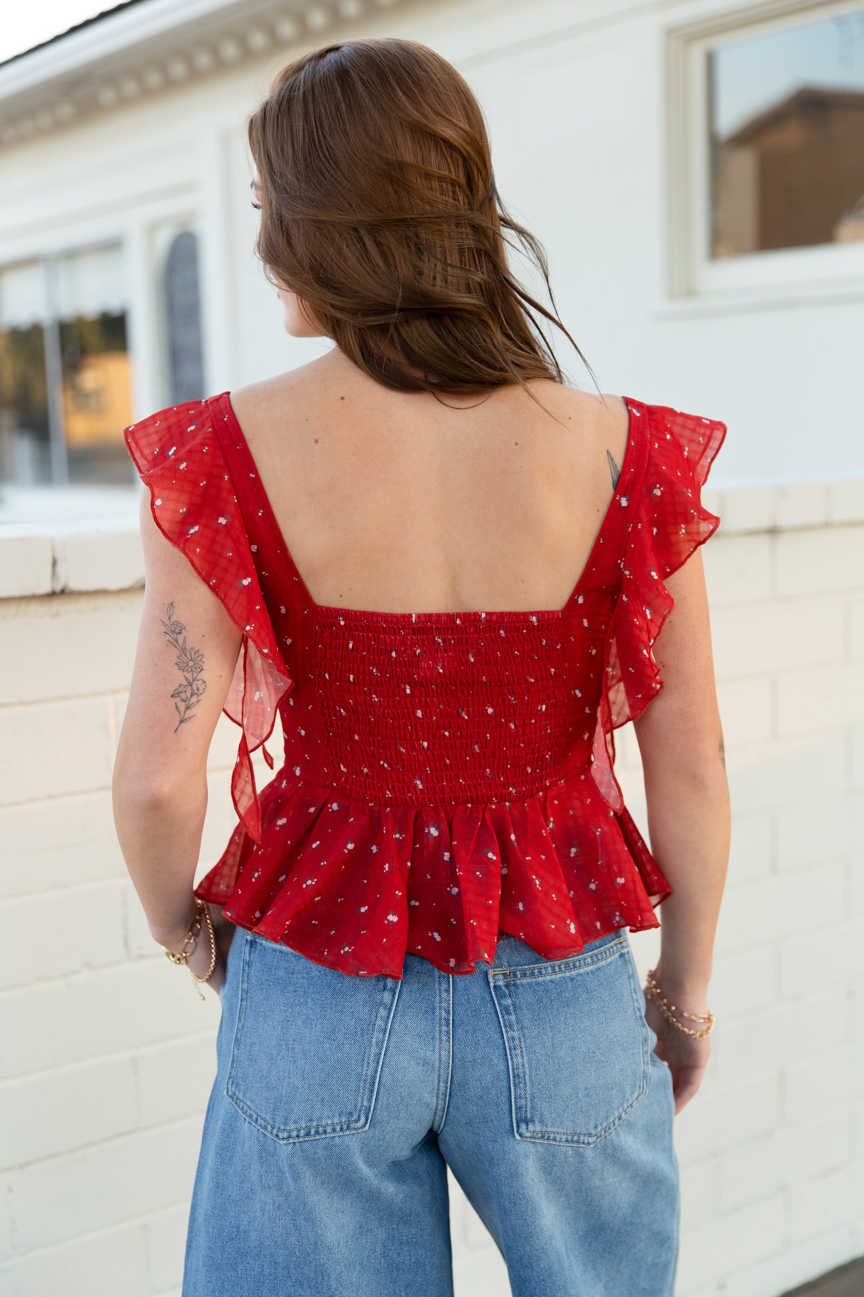 Woman wearing a red top with ruffled sleeves and blue jeans, standing against a white wall.