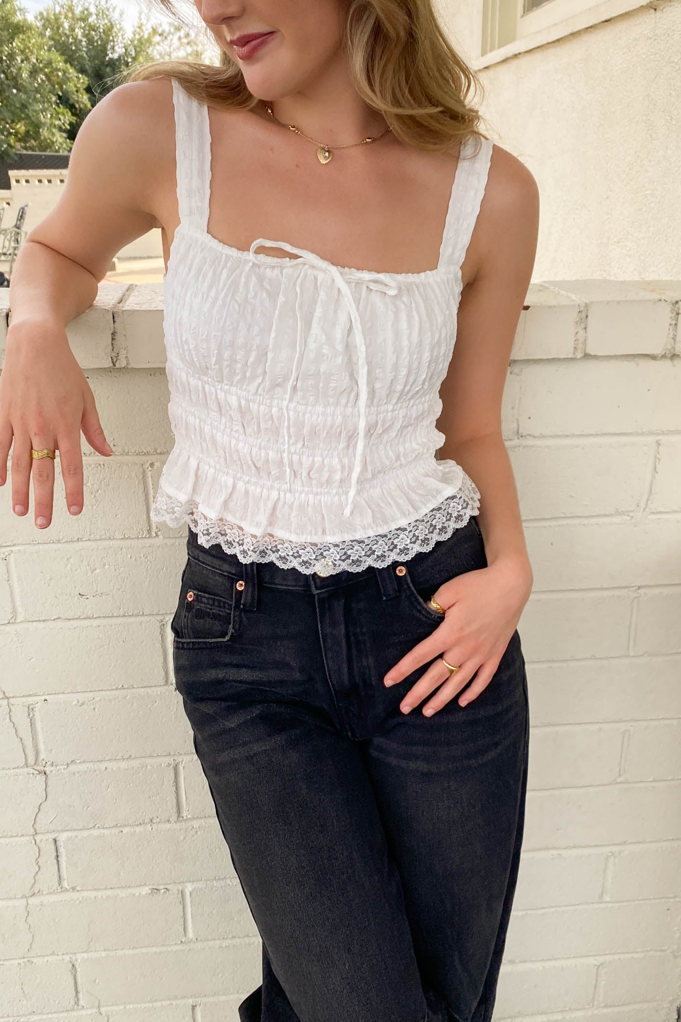 Woman wearing a white lace top and black jeans against a light brick wall.
