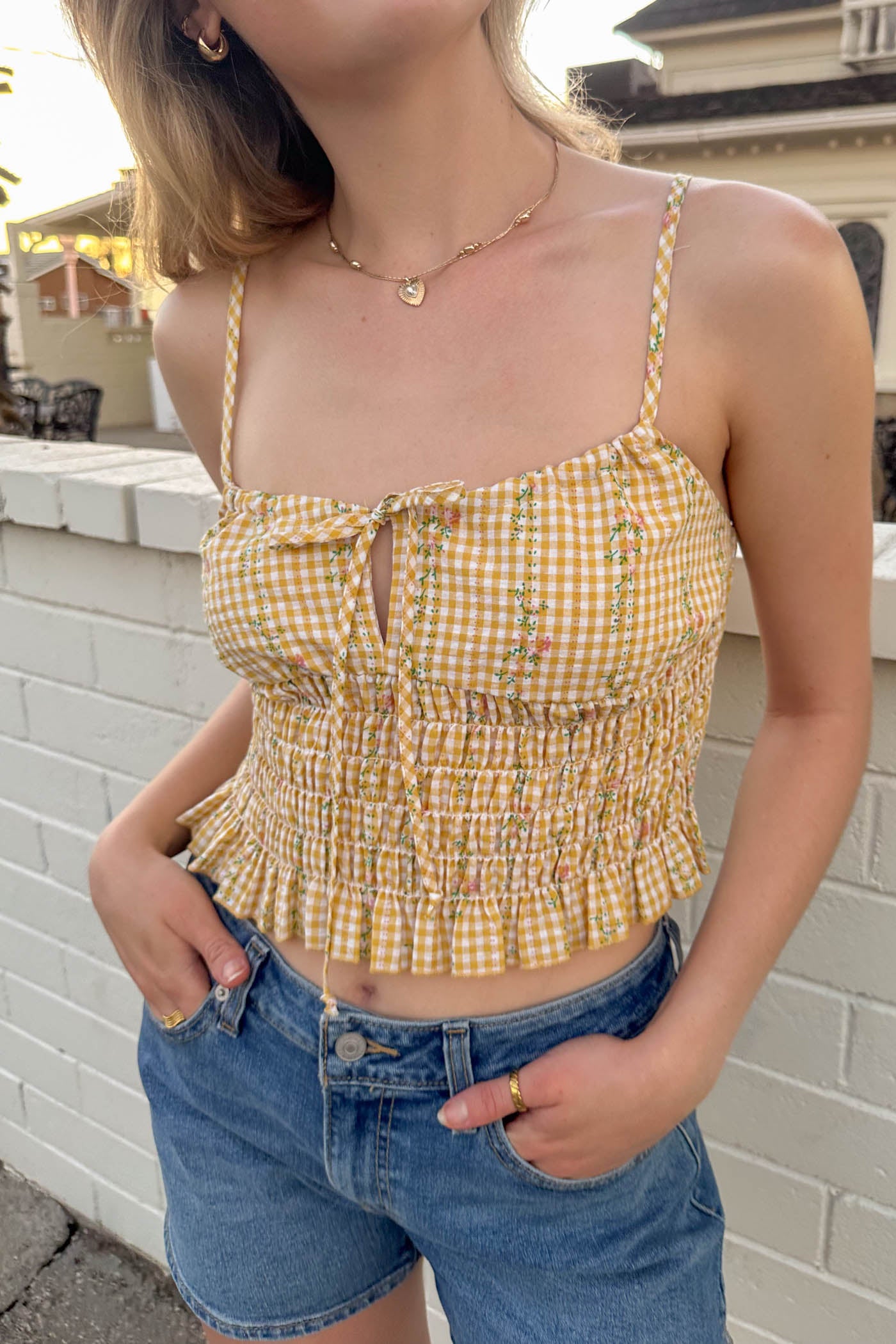 Woman wearing a yellow checkered top and blue shorts standing against a brick wall.