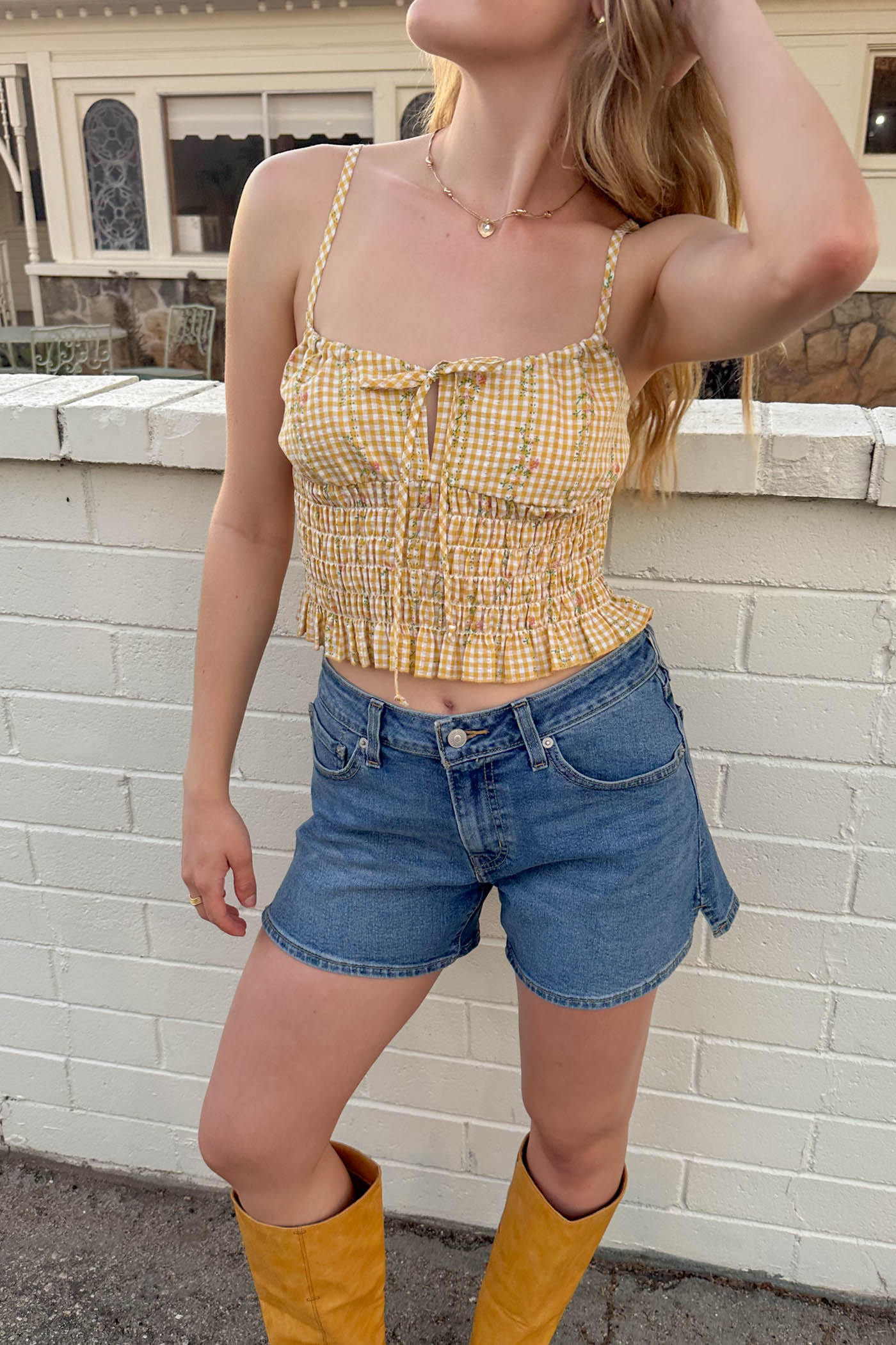 Woman wearing a yellow checkered top and denim shorts with yellow boots against a white brick wall.