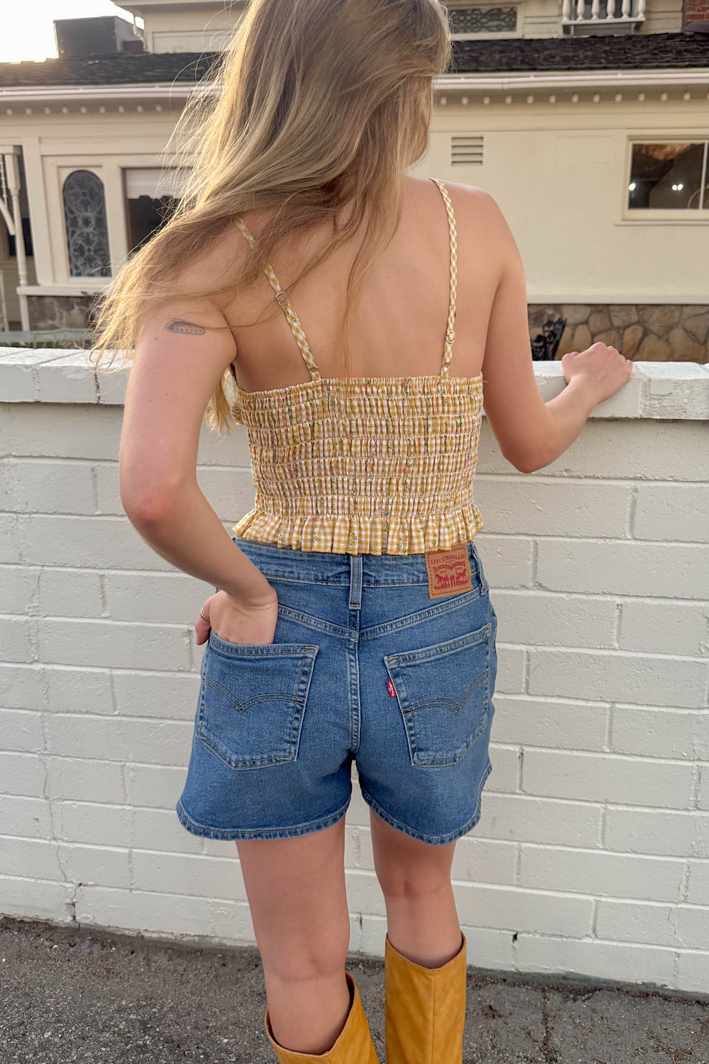 Woman wearing a yellow top and blue shorts with mustard boots standing against a white brick wall.