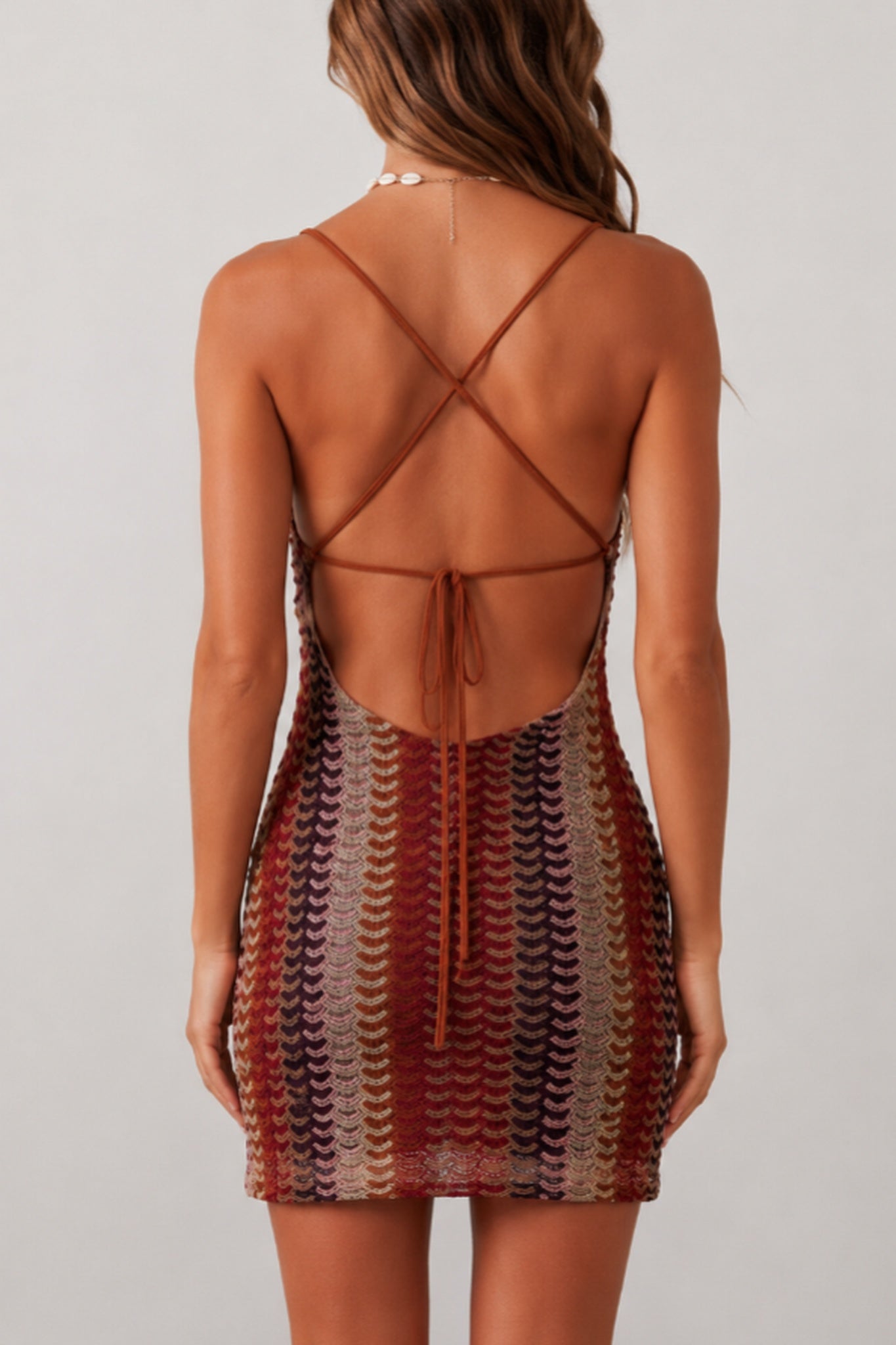 Rear view of a woman wearing a strappy open-back mini dress with a scalloped knit pattern in rust red, brown, beige, and dark accents, against a light gray studio background.
