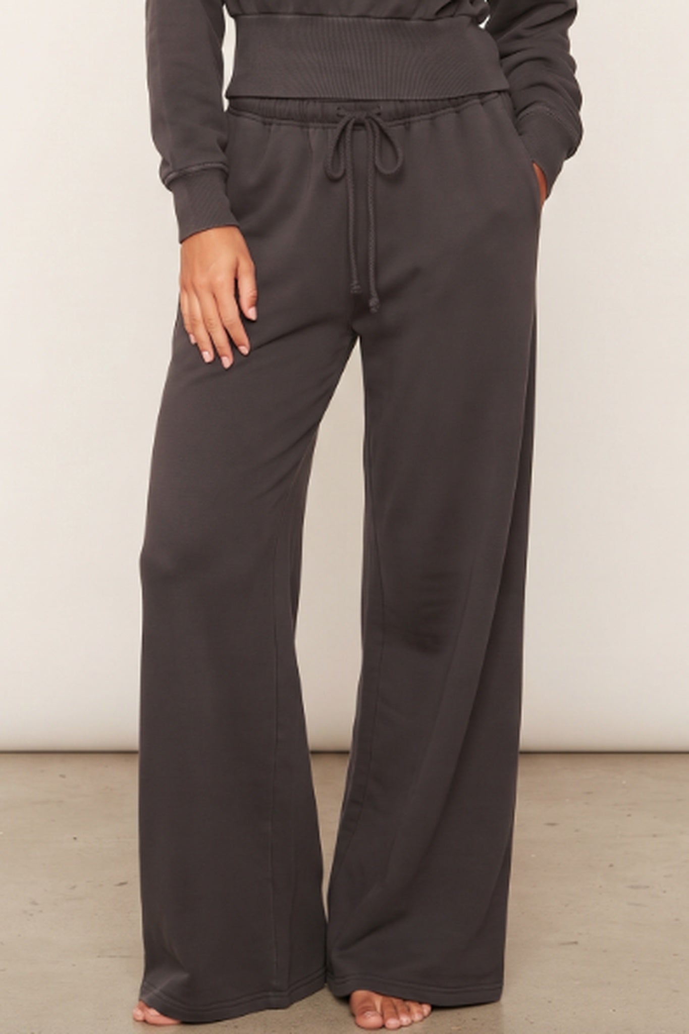 A person wearing dark charcoal wide-leg sweatpants with a drawstring waist and side pockets, paired with a matching dark sweatshirt, shown from the waist down against a plain light wall.