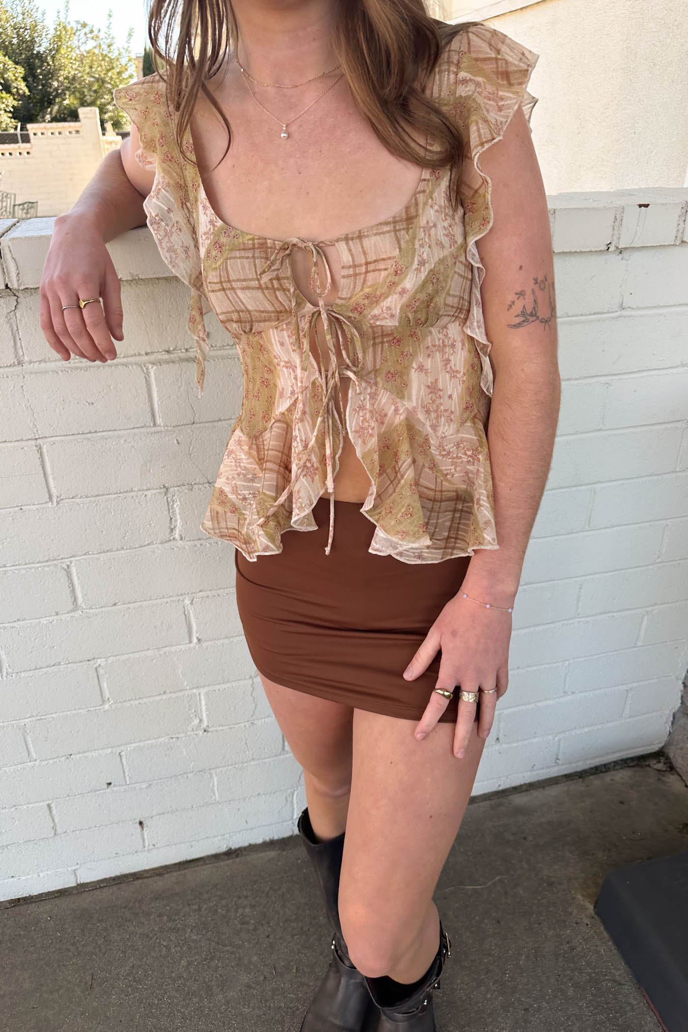 Woman wearing a layered outfit with a plaid top and brown skirt against a white brick wall.