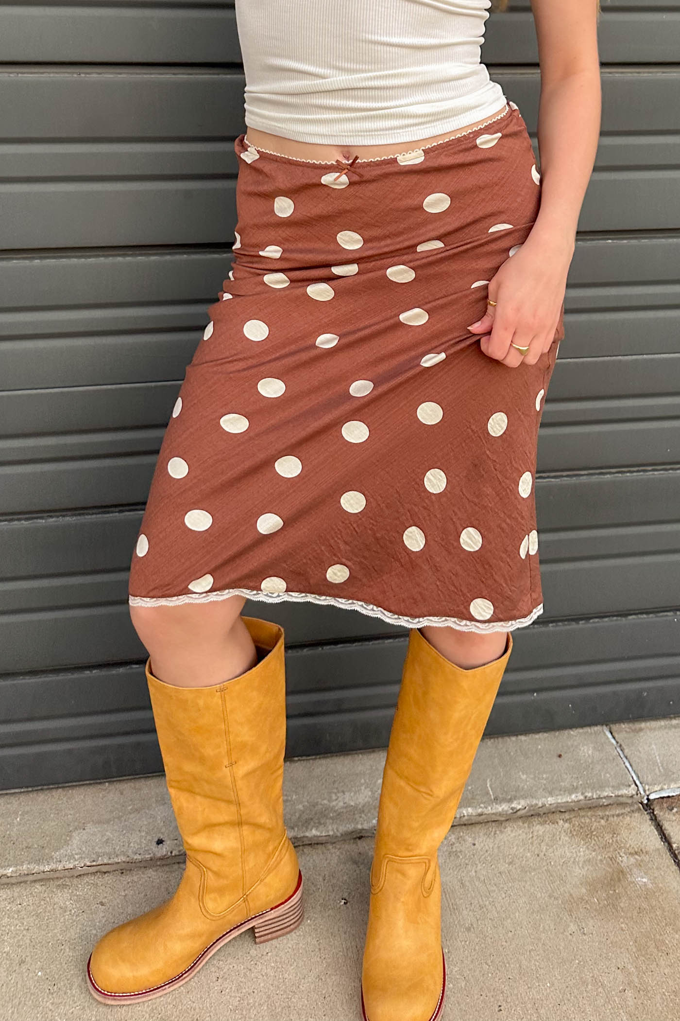 Person wearing a brown polka dot skirt and yellow boots against a gray wall.