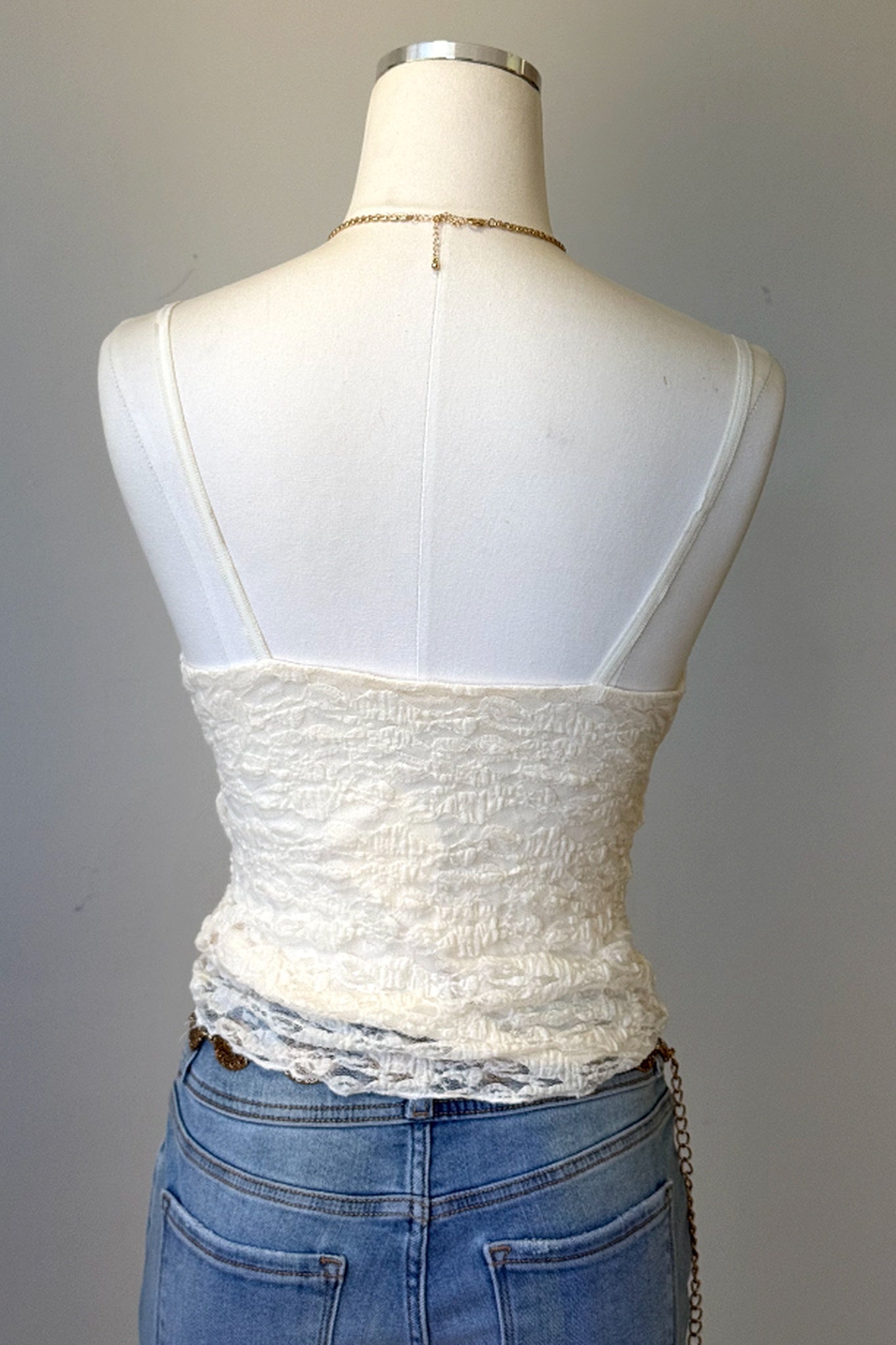 An off-white lace and smocked corset-style camisole top displayed on a white mannequin torso, paired with blue jeans against a plain gray background.