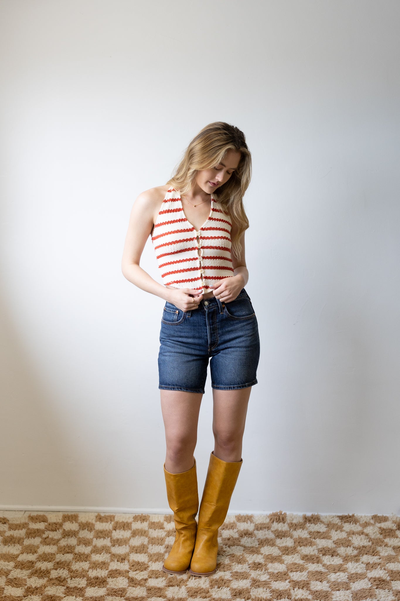 Woman wearing a striped tank top, denim shorts, and yellow boots against a plain wall.