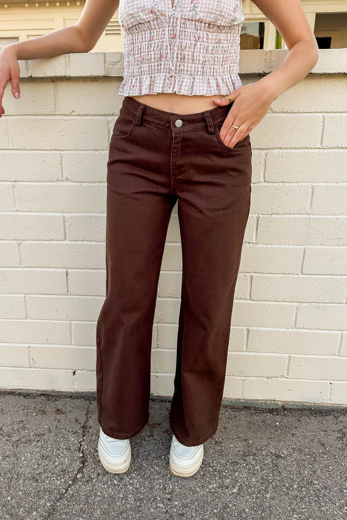 Person wearing brown pants and a patterned top against a beige brick wall.