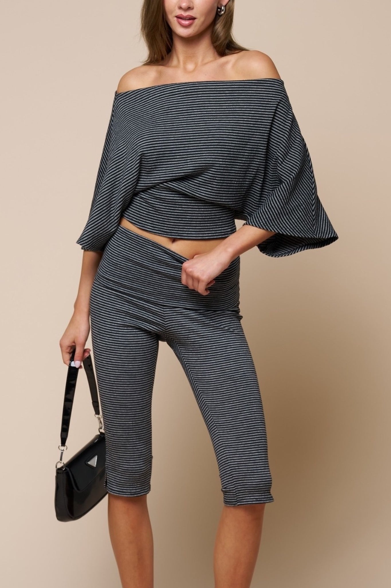 A woman modeling an off-the-shoulder two-piece set consisting of a dark gray and white striped crop top with flared bell sleeves and matching high-waisted striped capri pants, holding a black handbag against a solid beige background.