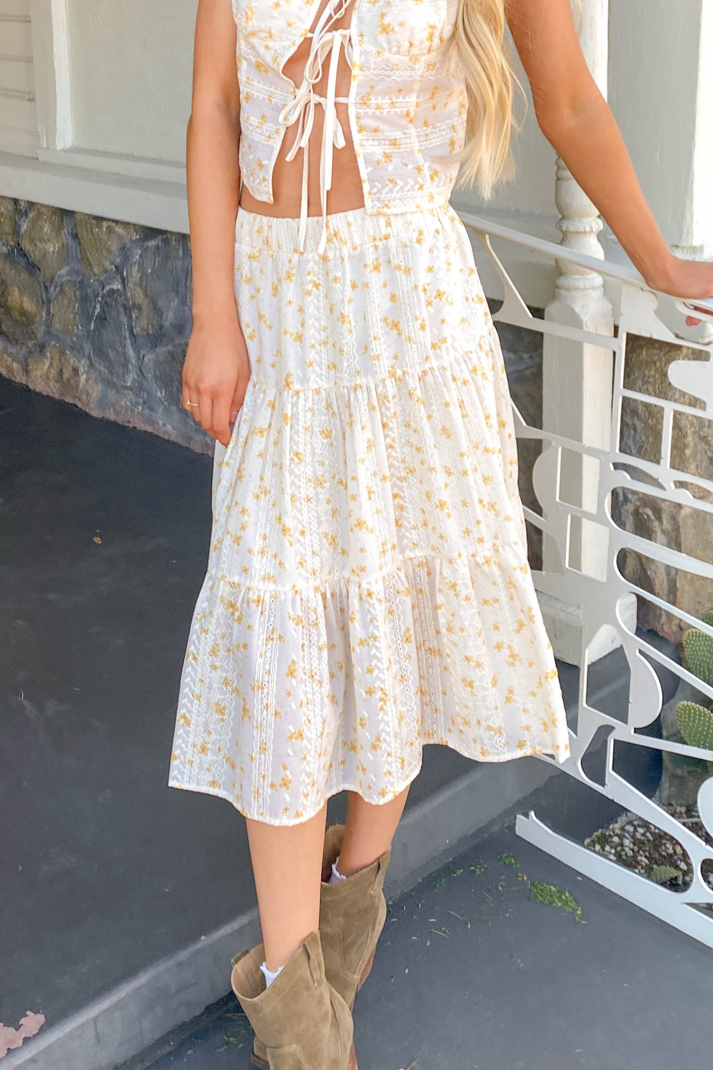 Woman wearing a white floral dress with cut-out shoulders and brown boots on a stone porch.