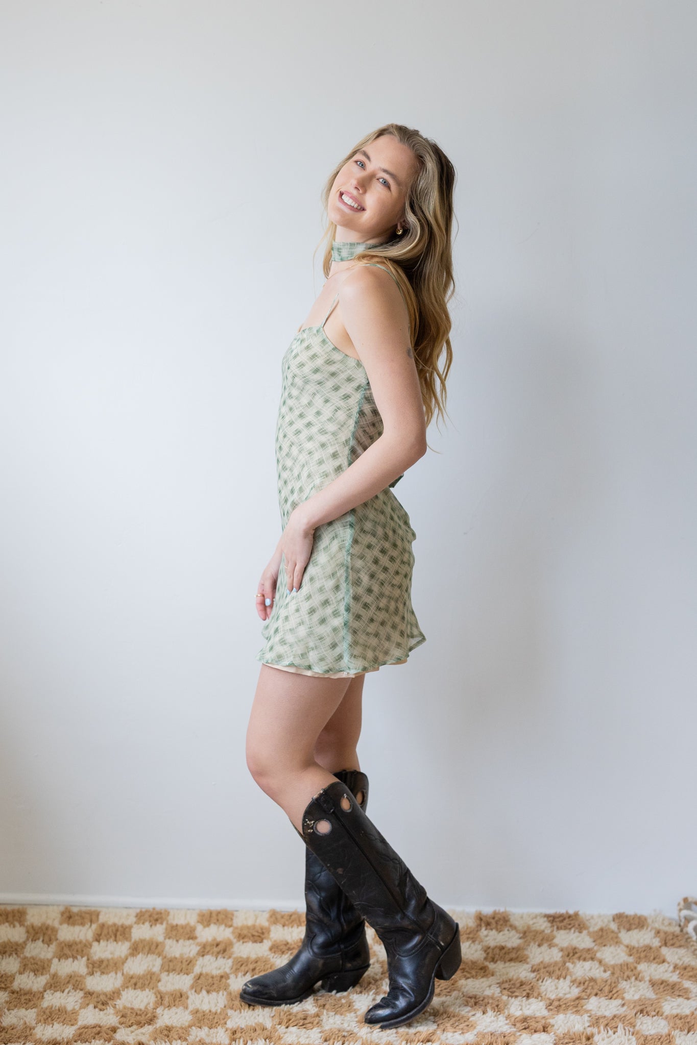 Woman wearing a patterned dress and black boots standing against a plain background