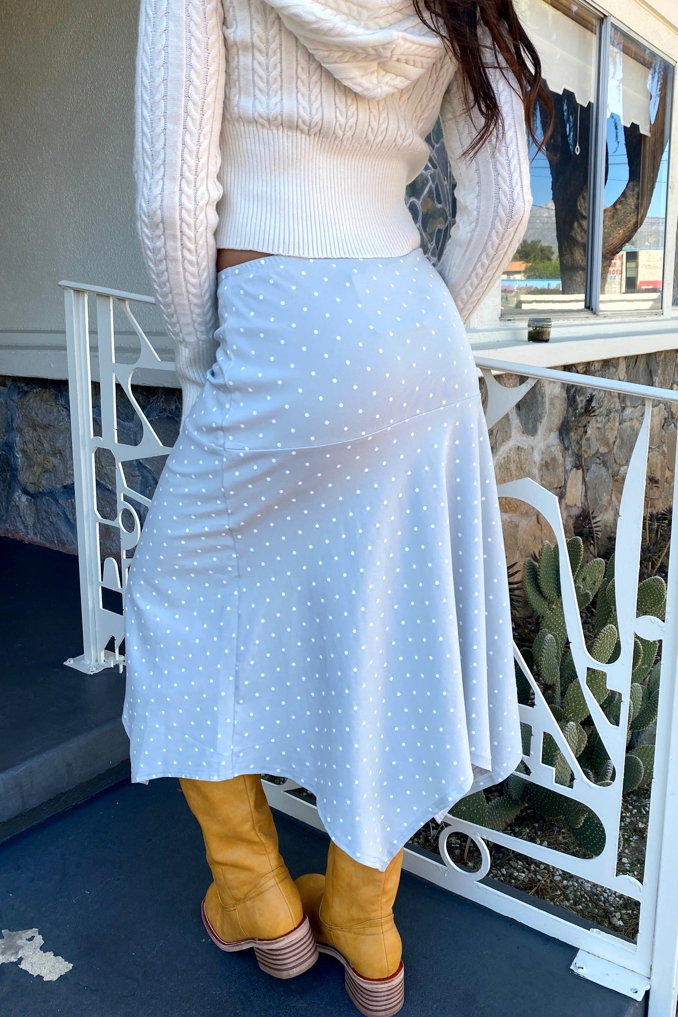 Person wearing a light blue polka dot skirt with a white sweater and yellow boots on a balcony.