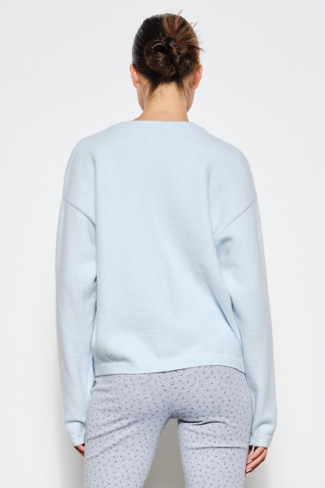 A woman viewed from behind wearing a light blue crewneck sweatshirt and gray patterned pajama pants against a plain white background.