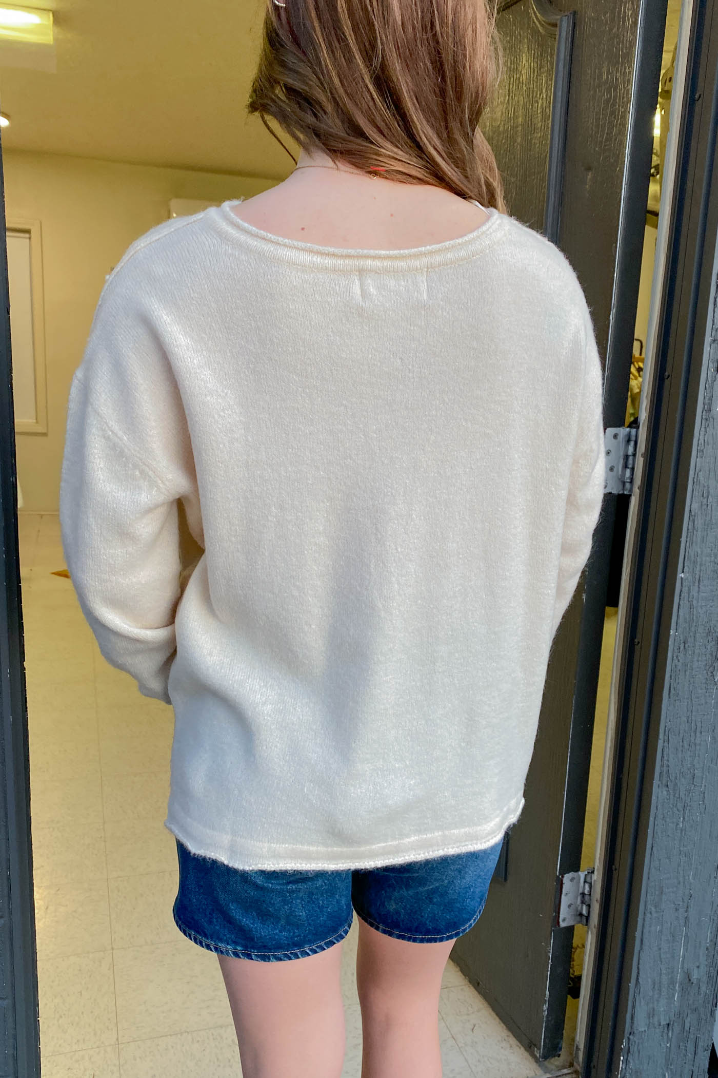 Person wearing a white sweater and blue shorts standing in a doorway.
