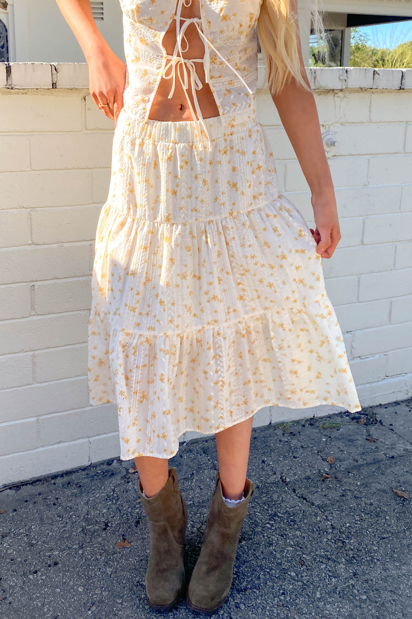 Person wearing a white floral dress with an open back and brown boots standing against a white brick wall.