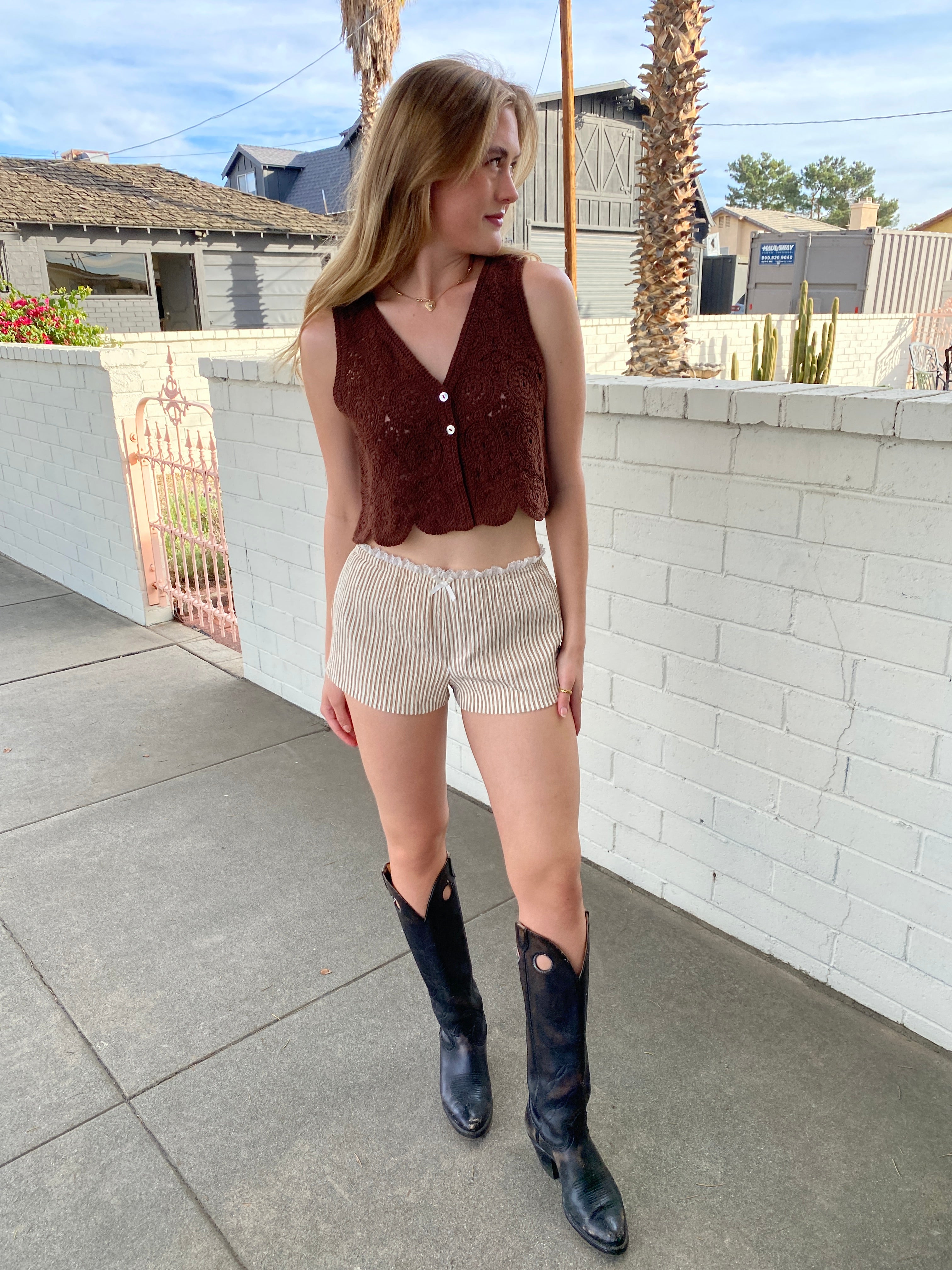 Person standing on a sidewalk wearing a brown top, beige shorts, and black boots.
