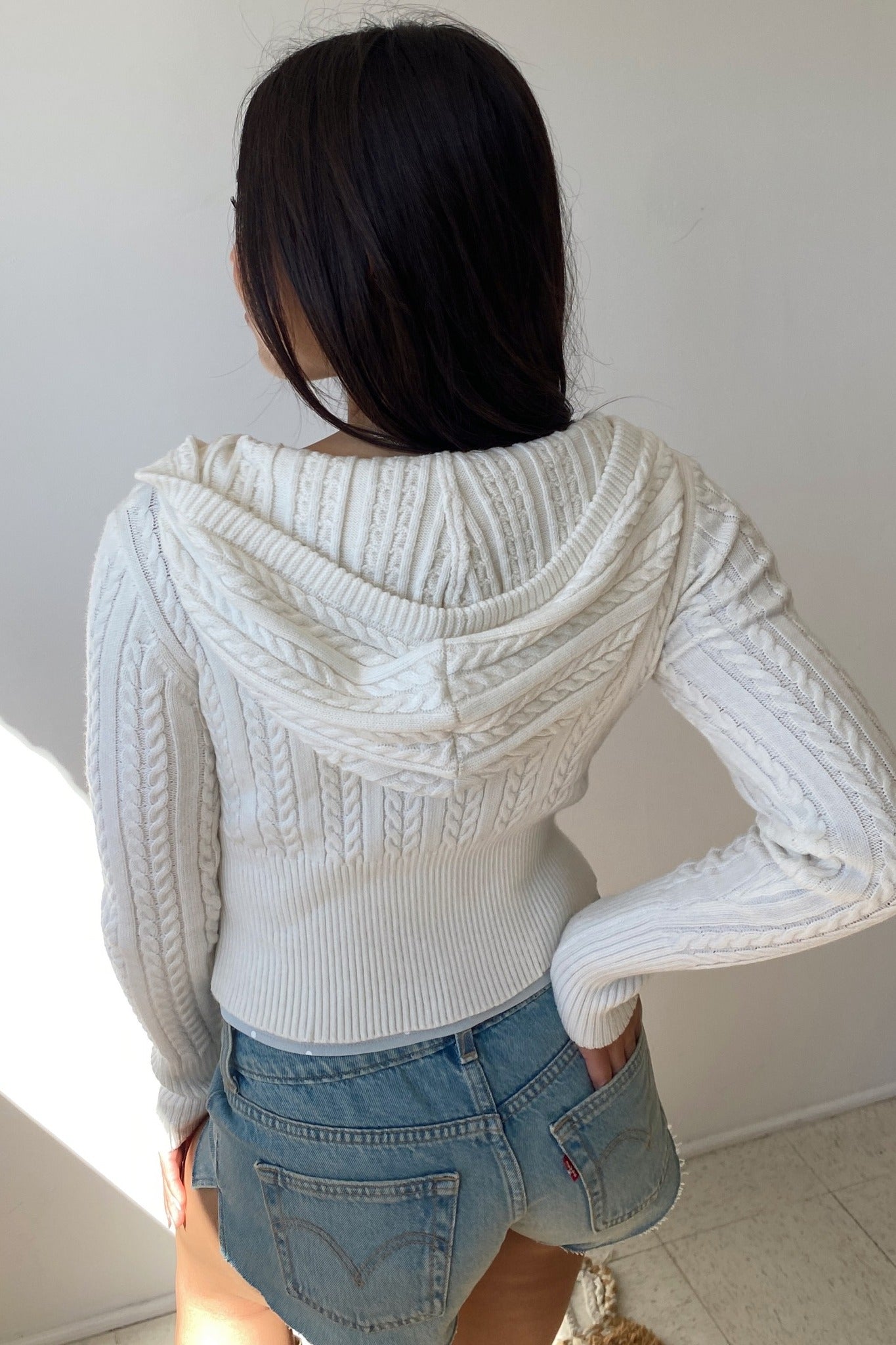 Person wearing a white cable knit hoodie and denim shorts against a plain background