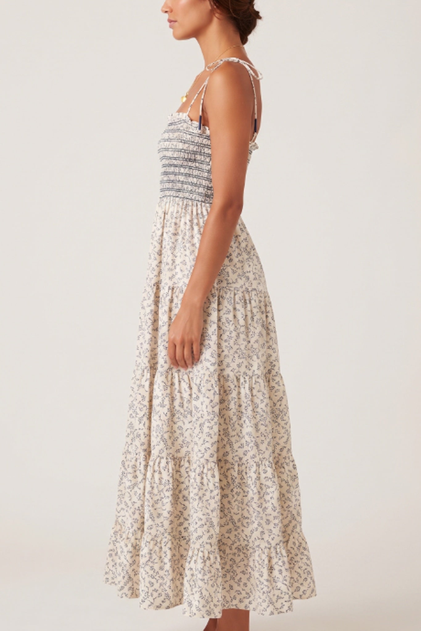 A woman wearing a sleeveless tiered maxi dress with a smocked bodice and a small floral print, shown from the side against a plain light background.
