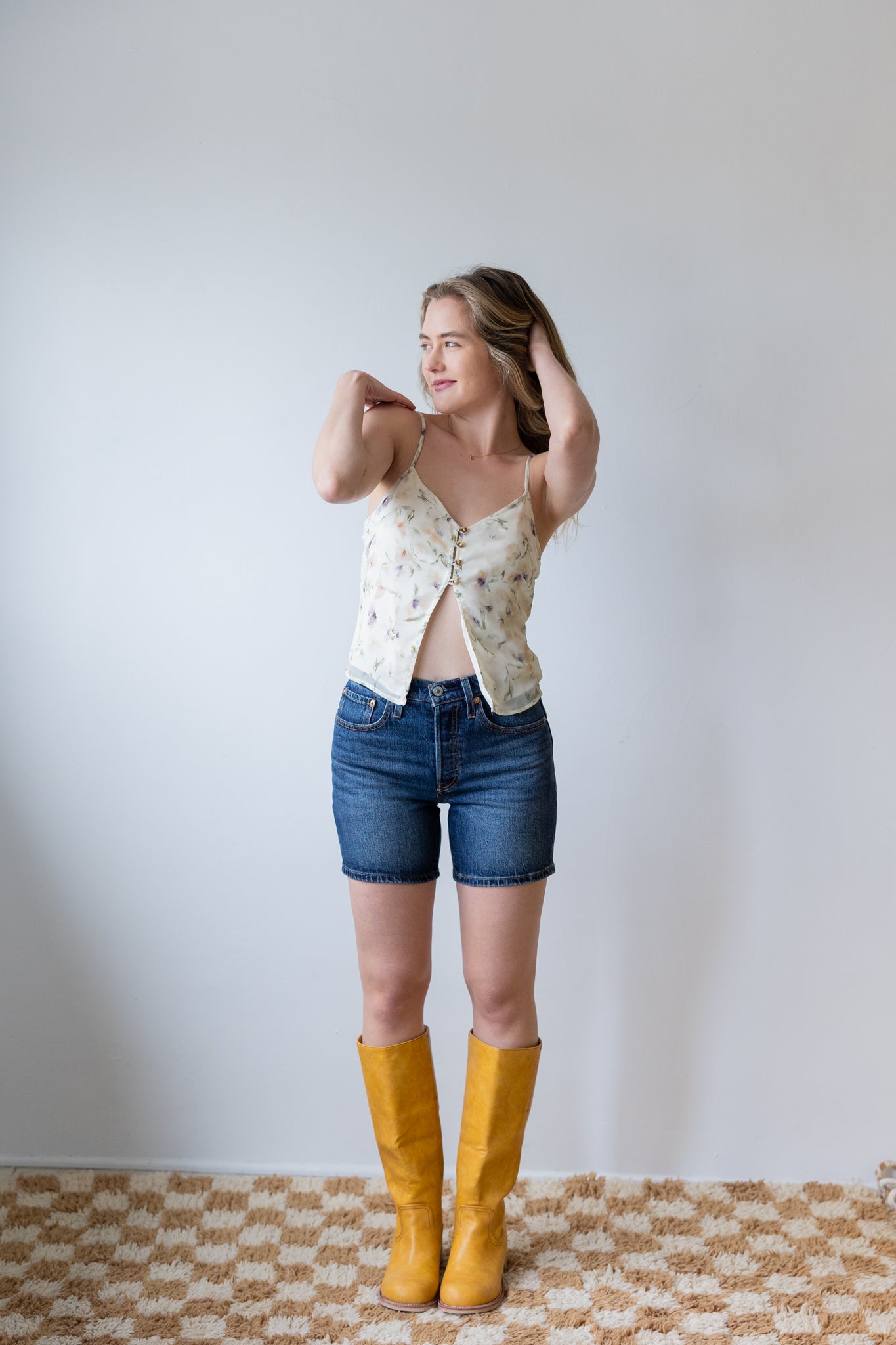 Woman wearing a patterned top, denim shorts, and yellow boots against a plain background