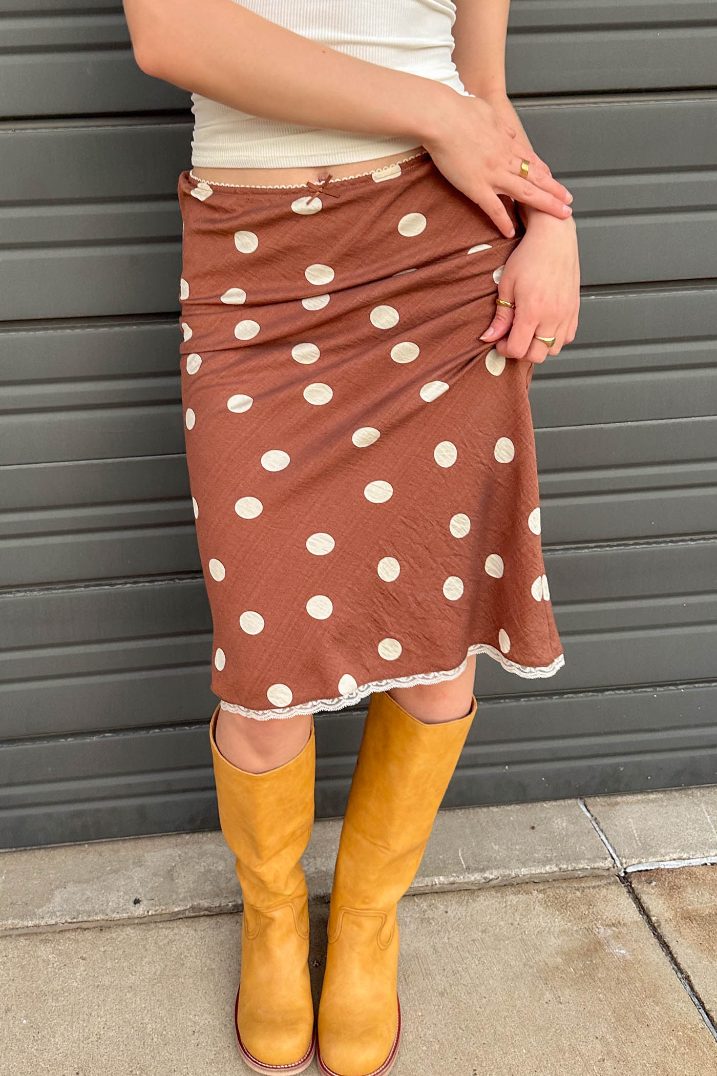 Person wearing a brown polka dot skirt and yellow boots against a gray wall.