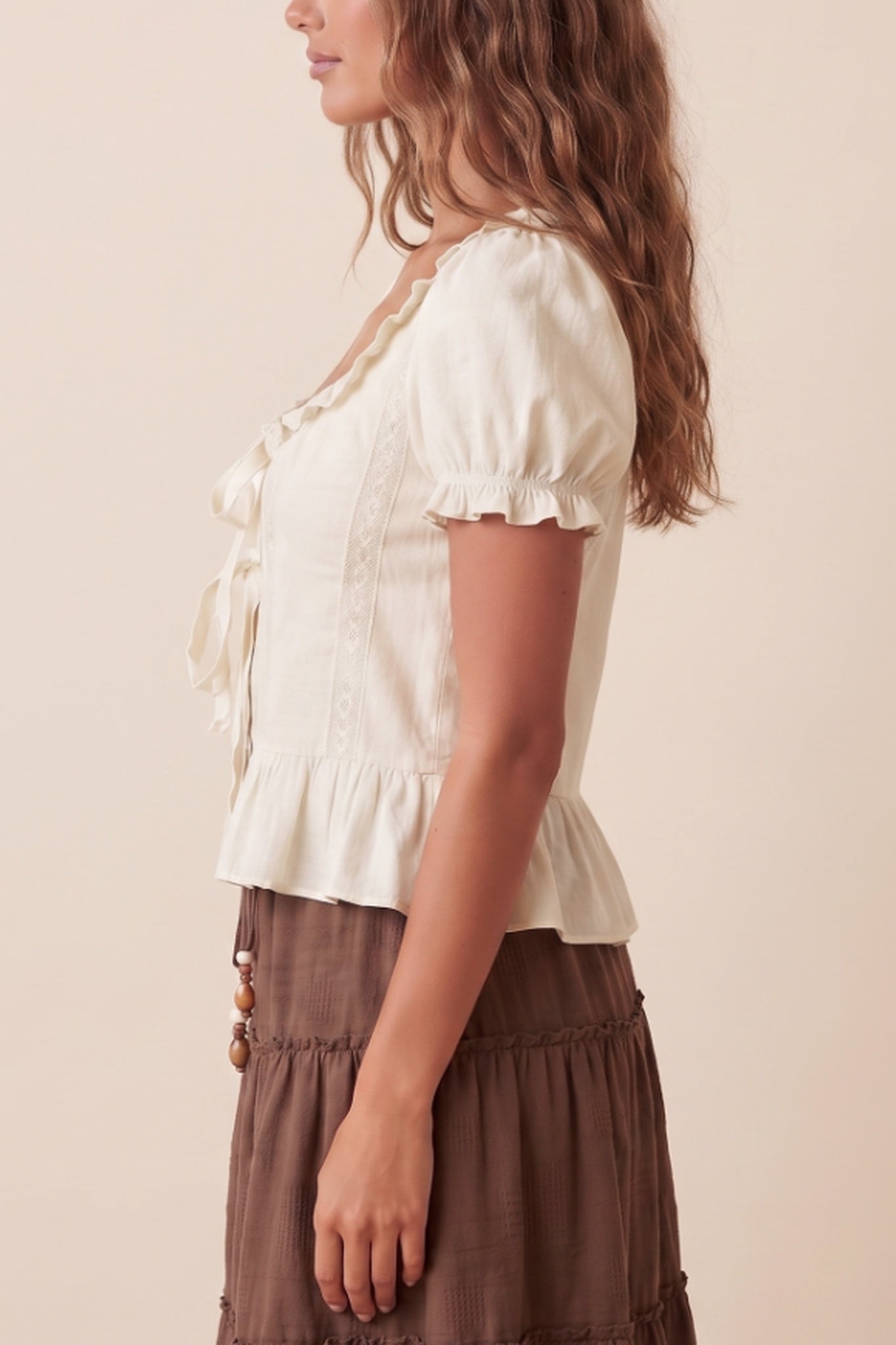 A woman shown in side profile wearing a cream puff-sleeve blouse with a ruffled neckline and tie detail, paired with a brown skirt, against a plain beige studio background.