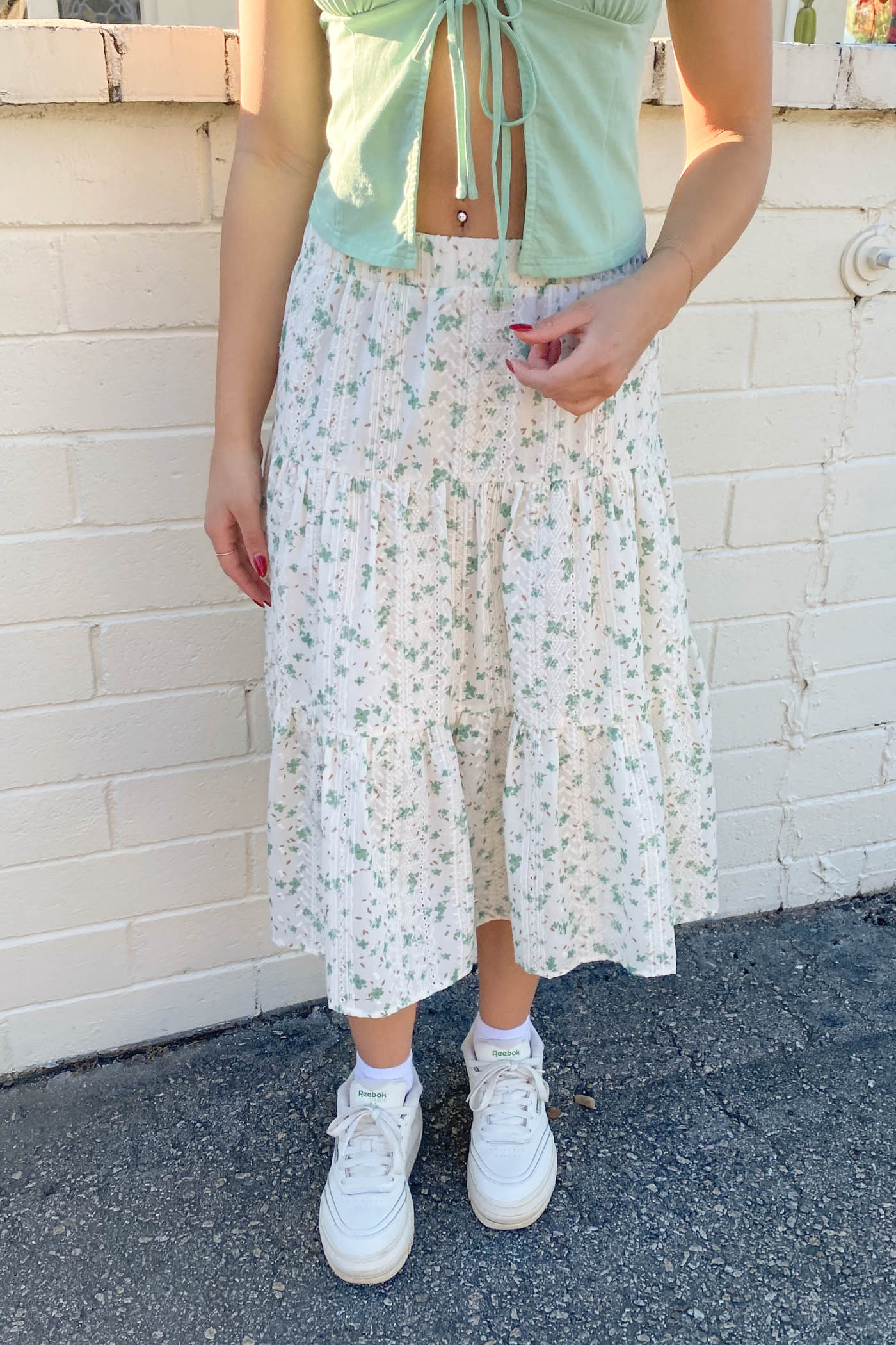 Person wearing a light green top and floral skirt with white sneakers against a white brick wall.
