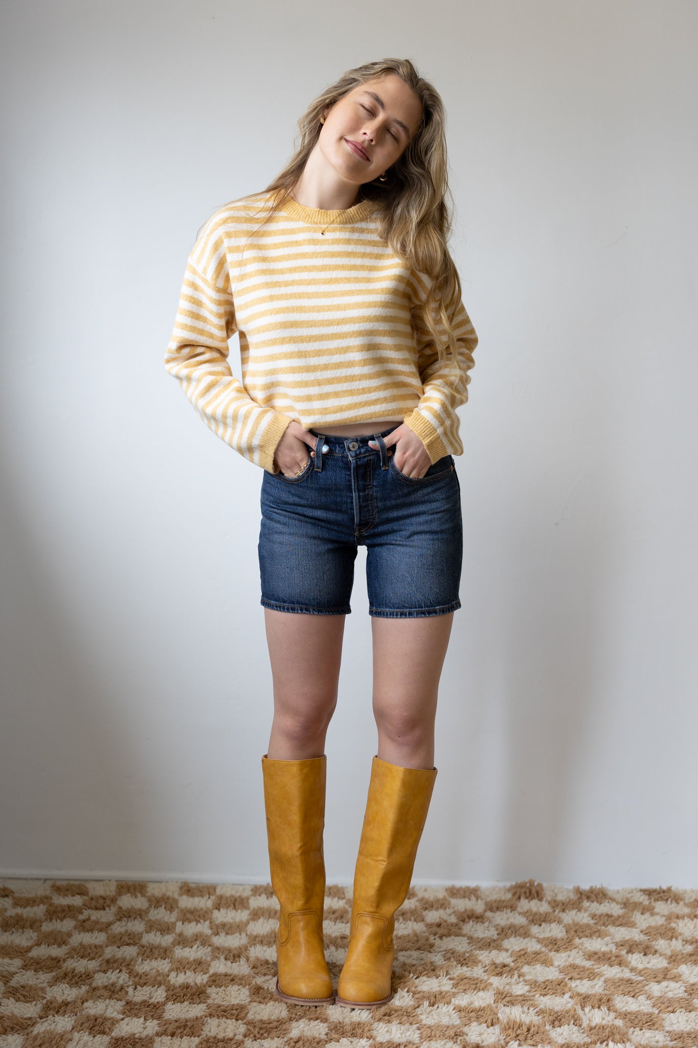 Woman wearing a yellow striped sweater, blue shorts, and yellow boots against a plain background