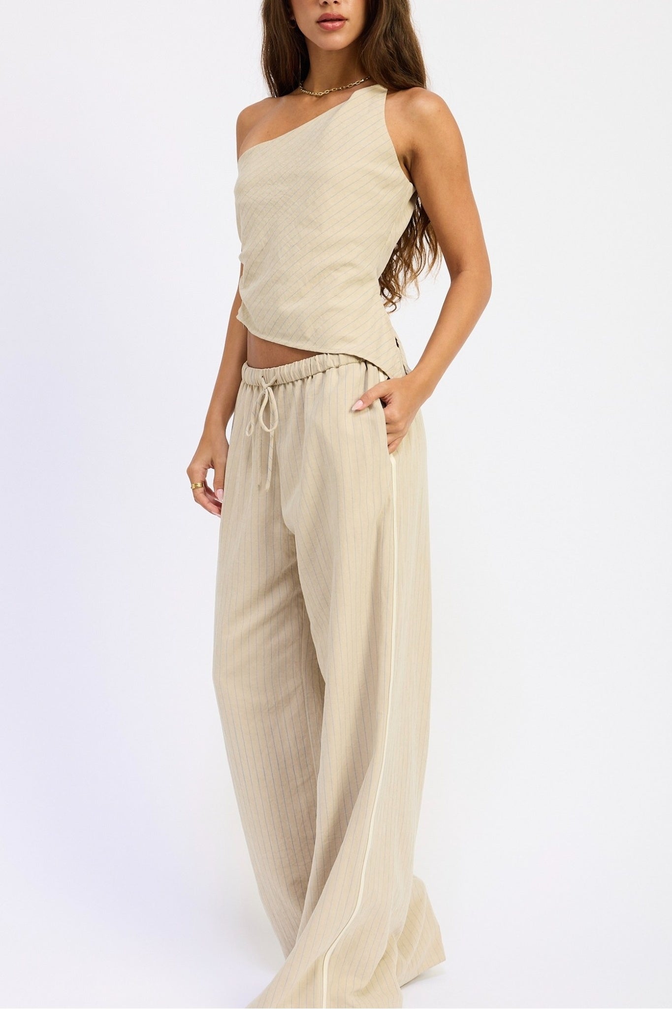 A woman modeling a light beige two-piece outfit consisting of a textured one-shoulder crop top and matching wide-leg drawstring pants, set against a plain white studio background.