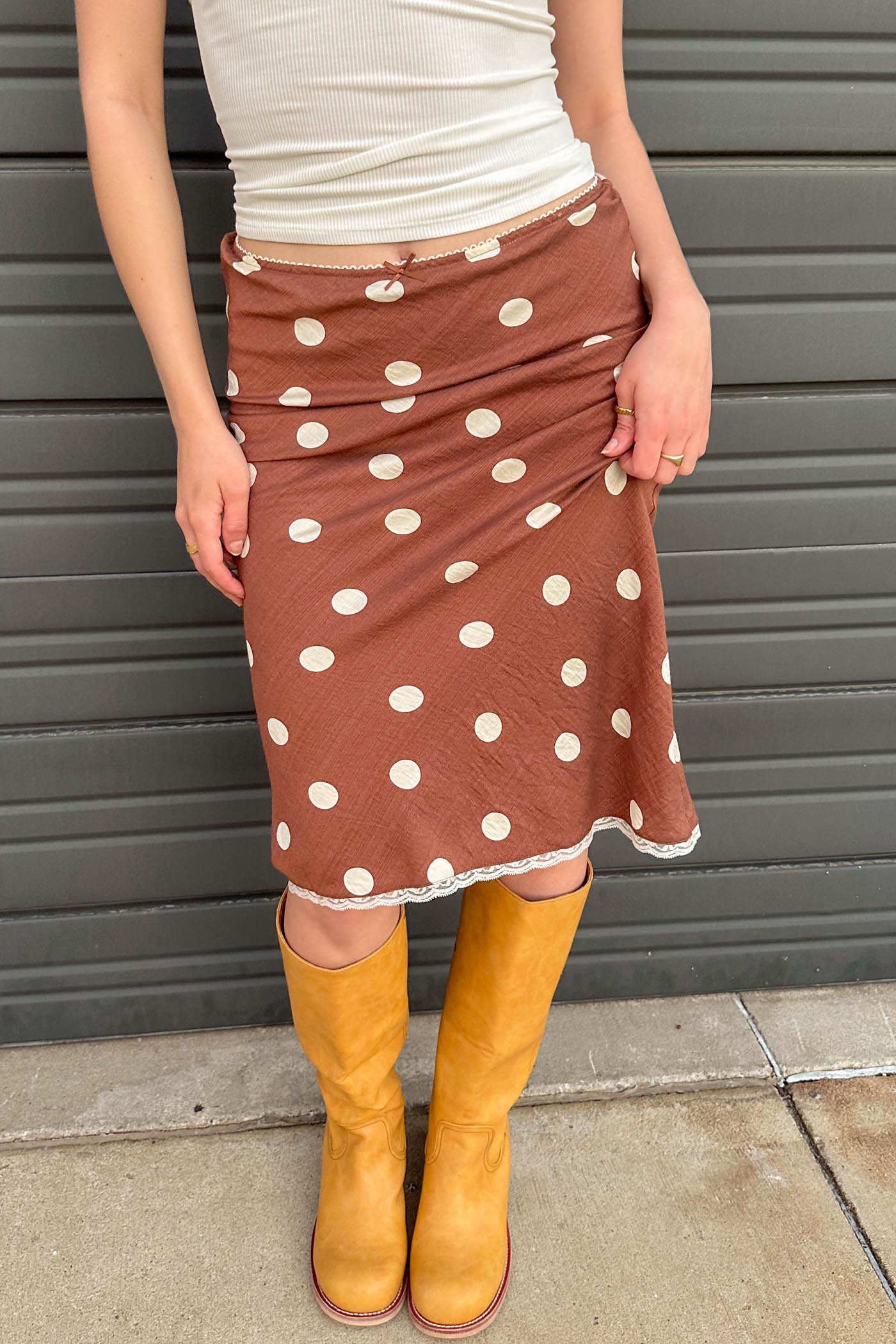 Person wearing a brown polka dot skirt and yellow boots against a gray wall.