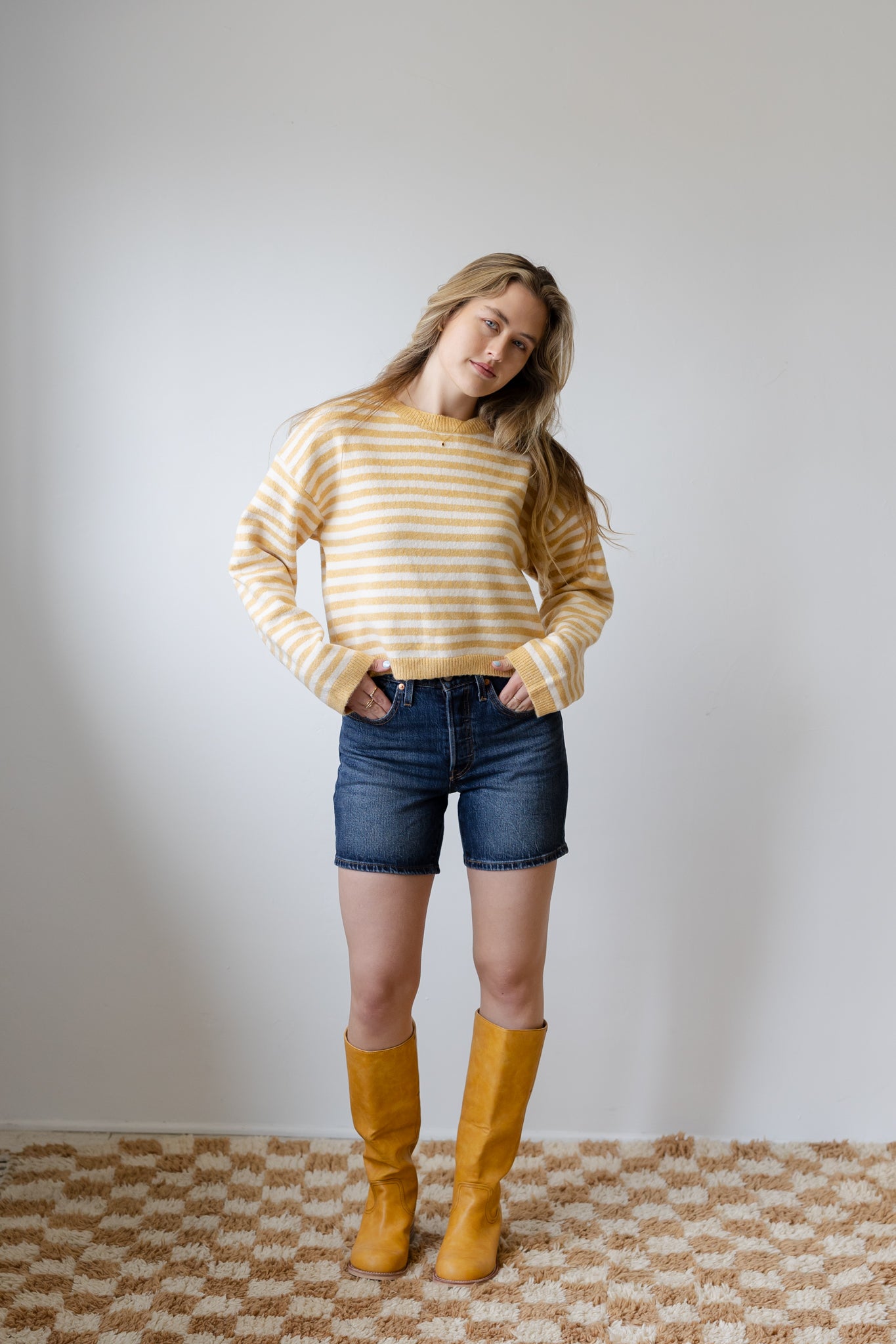 Woman wearing a striped sweater, denim shorts, and yellow boots against a plain background