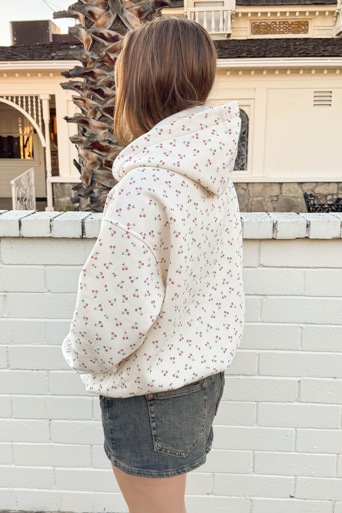 Person wearing a floral hoodie and denim shorts standing in front of a white brick wall.