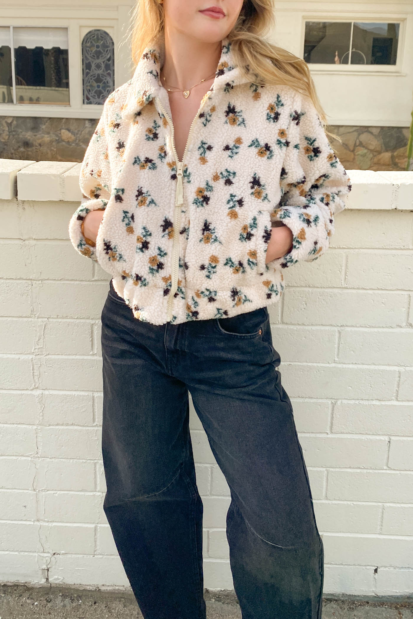 Person wearing a floral blouse and jeans standing against a white brick wall.